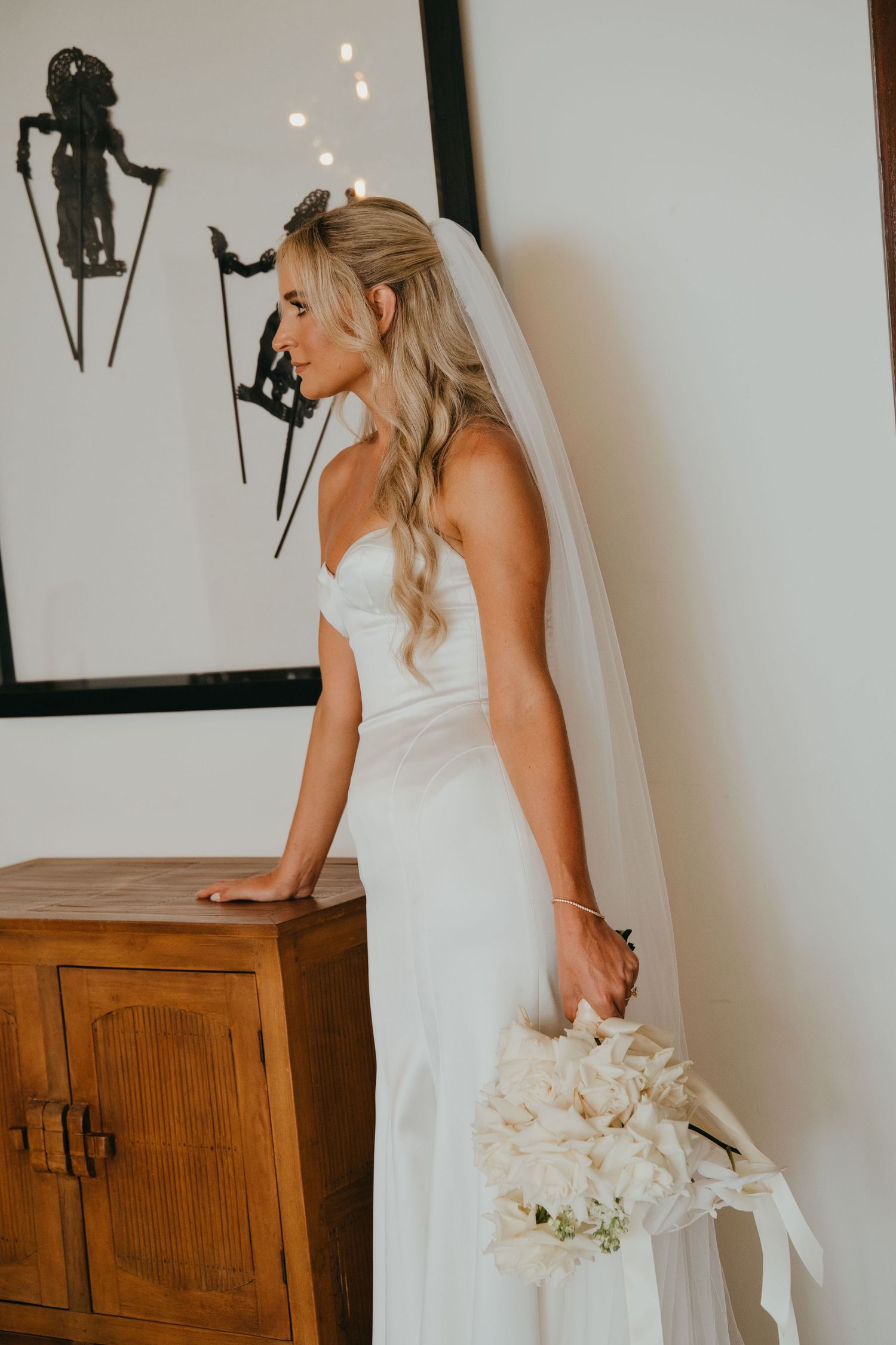 Elegant bride in a white gown holding a bouquet with a veil by a wooden cabinet.