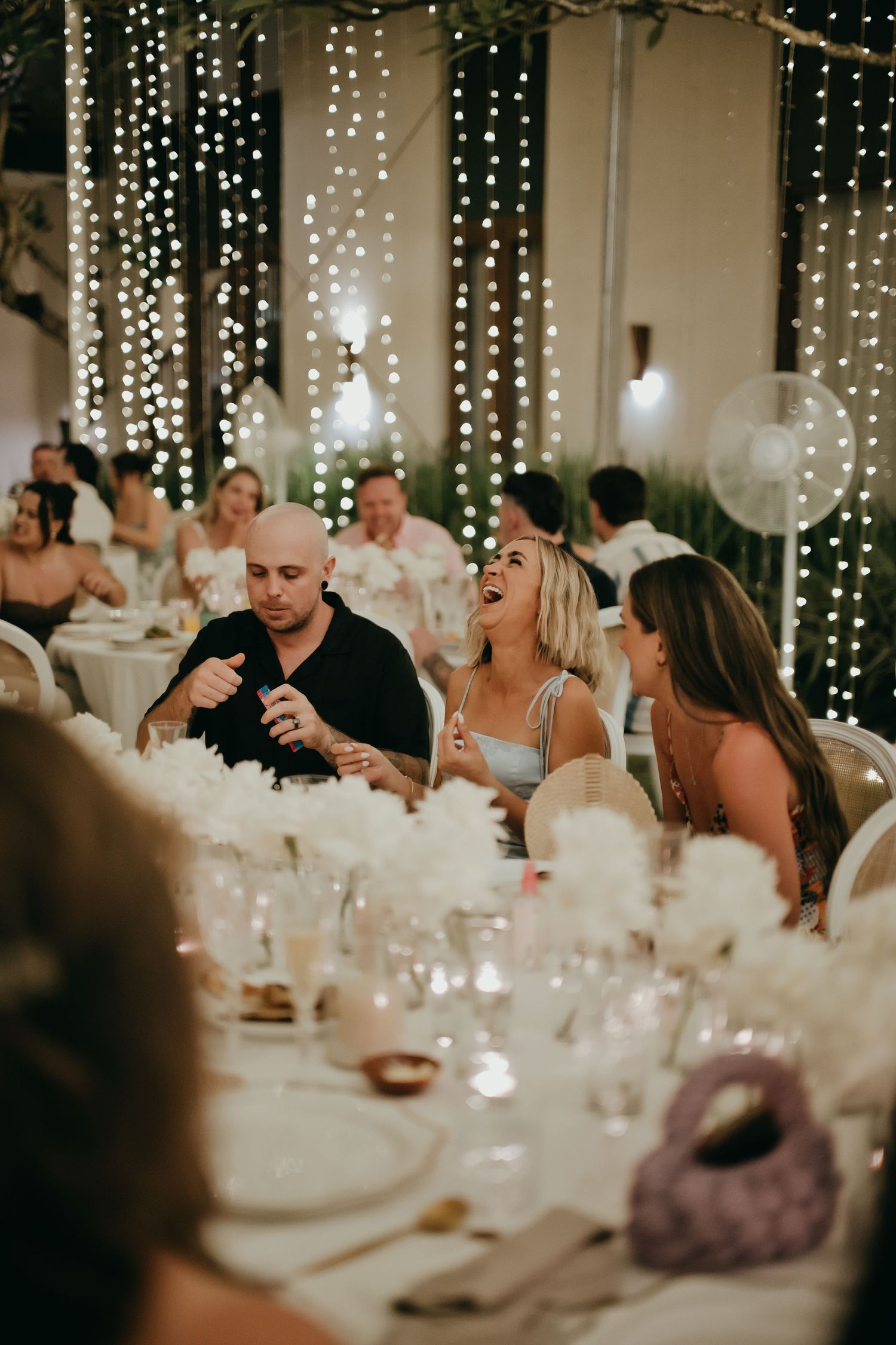 Wedding guests laugh and celebrate together under sparkling fairy lights at the reception dinner.