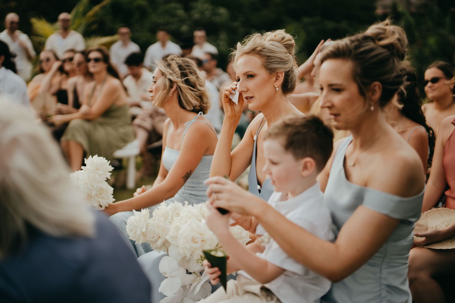 Emotional bridesmaids in blue dresses wiping tears during outdoor wedding ceremony.
