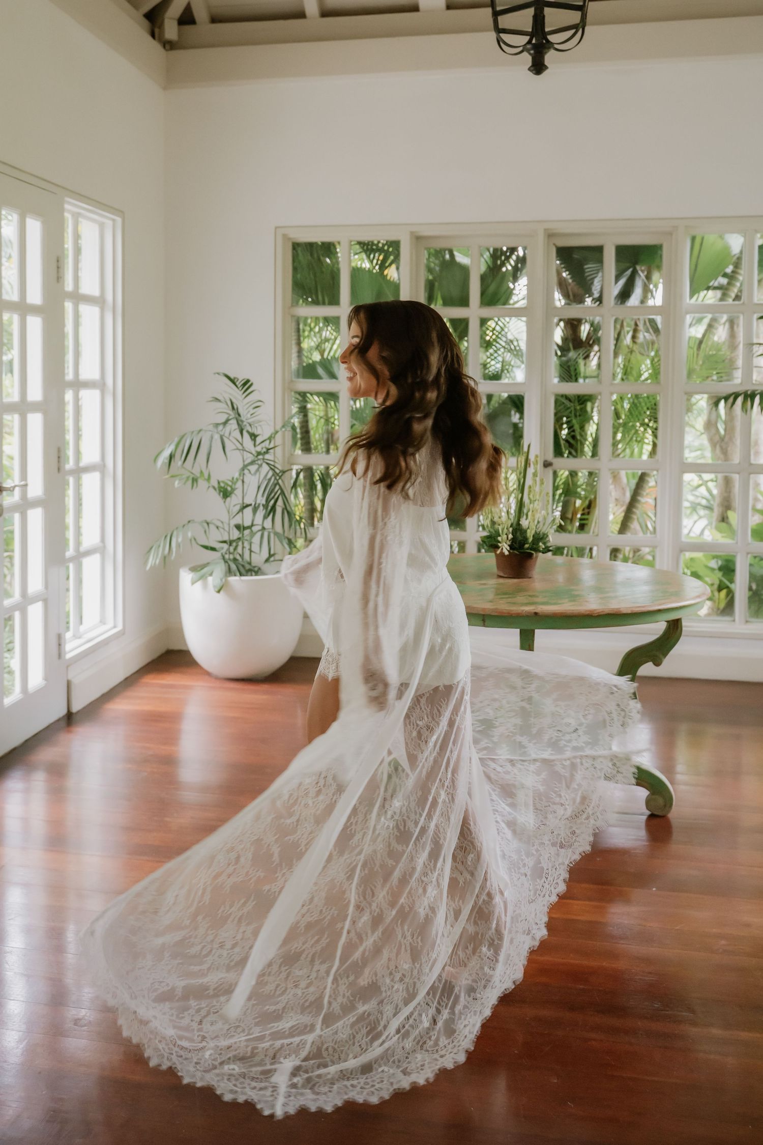 Bride twirling in a flowing white lace bridal robe in a bright tropical villa with hardwood floors.