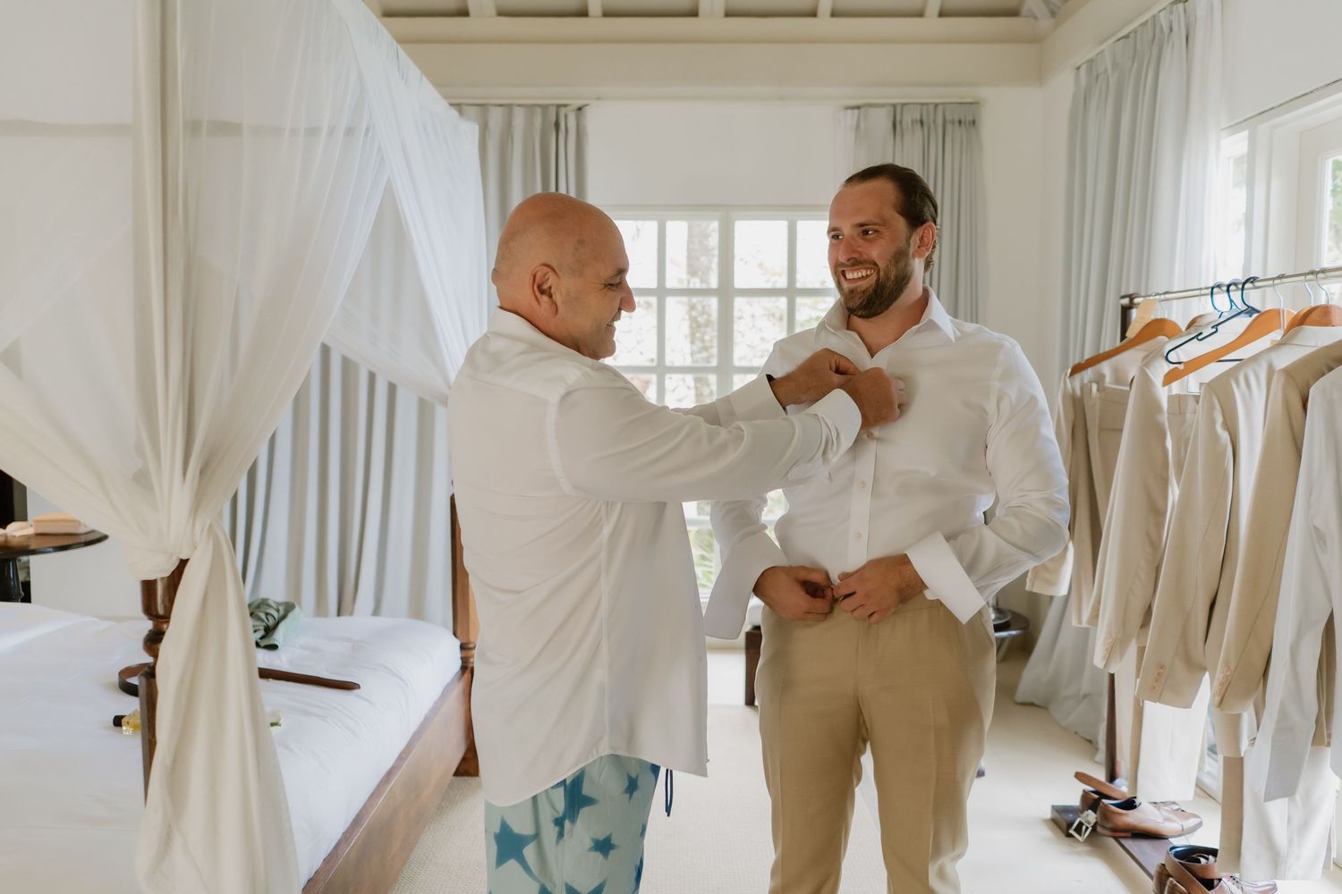 Two men in a bright room, one helping the other button a white shirt, with hanging suits in the background.