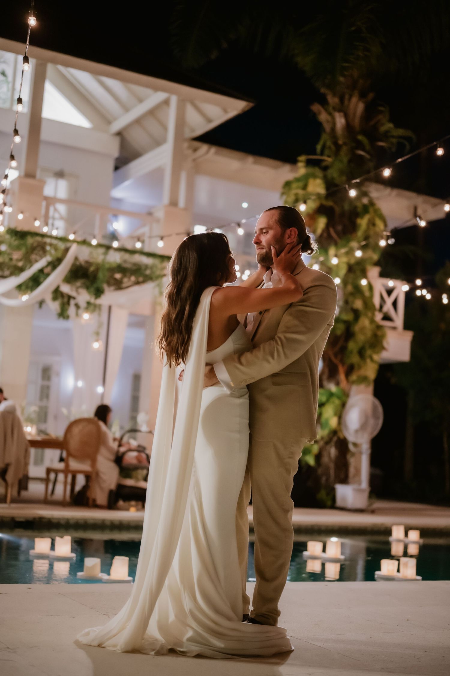 Newlyweds share romantic first dance beside candlelit pool with string lights and lush greenery.