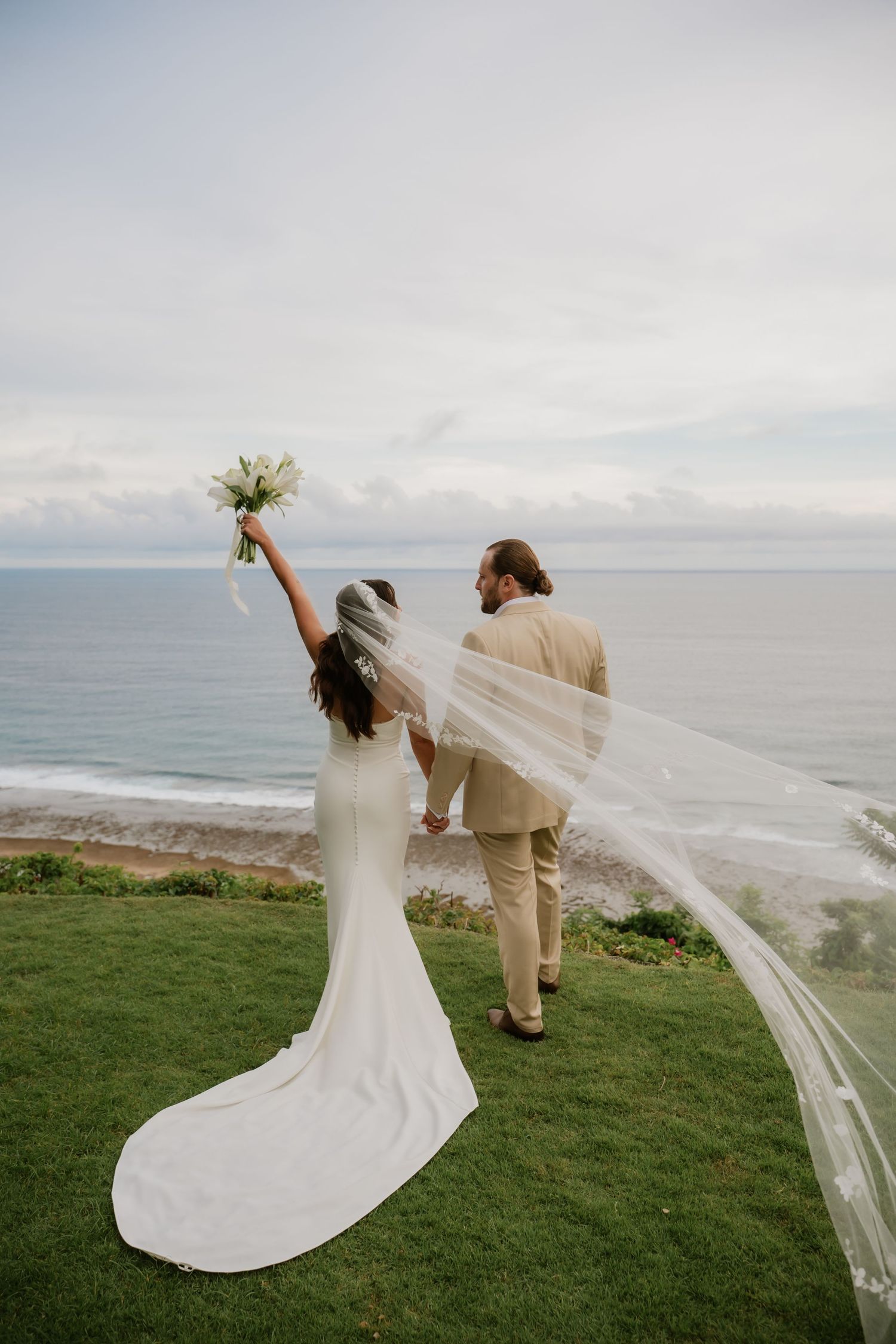Bride raises bouquet as couple stands on cliff overlooking ocean, veil flowing dramatically in wind.