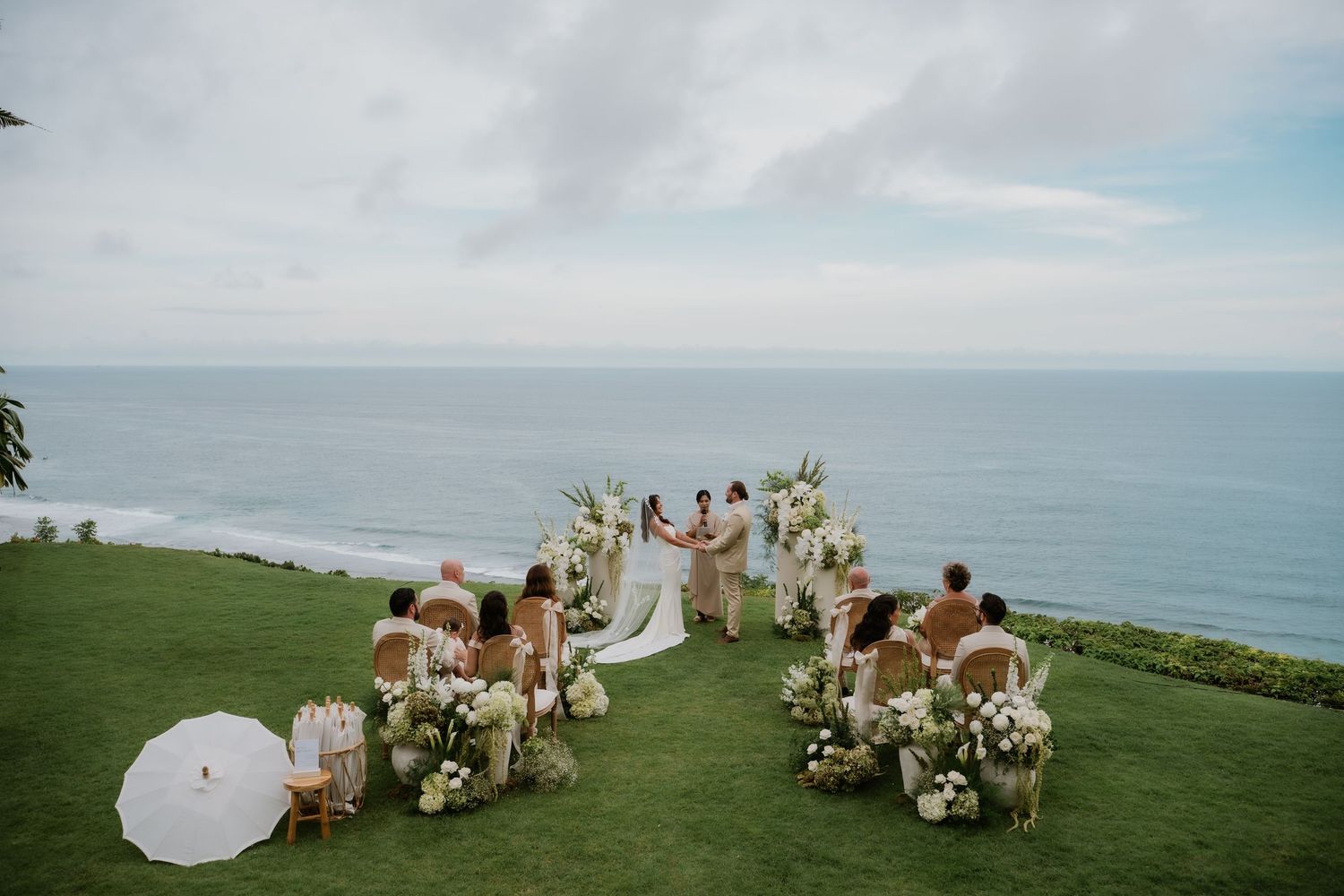 Intimate outdoor wedding ceremony on a cliffside overlooking the ocean with white floral decor.