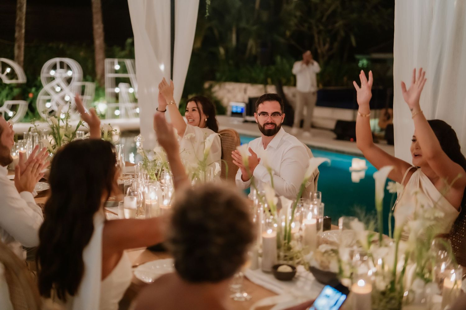 Wedding guests raise hands in celebration during lively outdoor reception dinner by the pool.