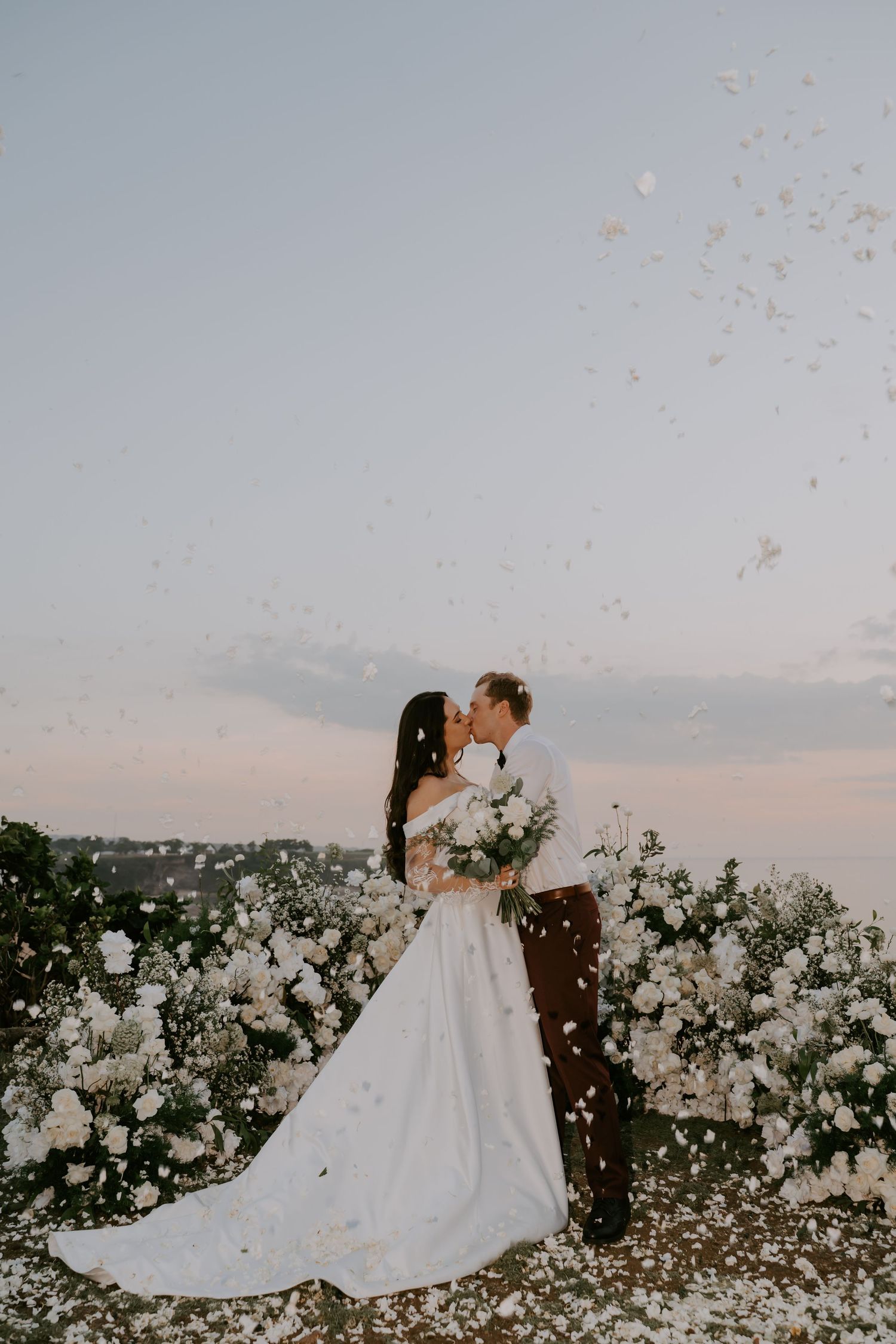 Bride and groom kiss surrounded by white flowers as confetti falls at sunset.
