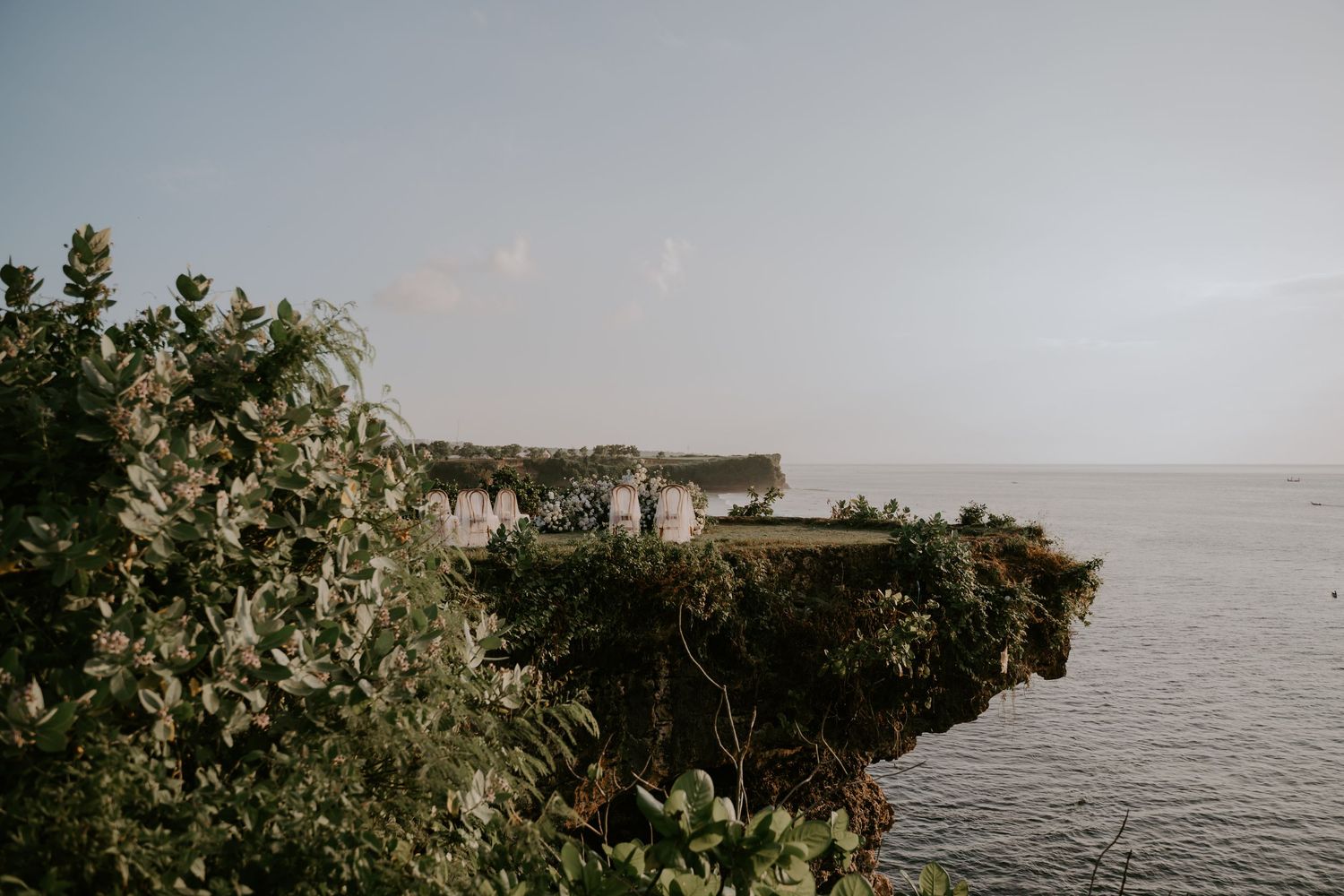 Lush green cliffside overlooking calm ocean waters under soft hazy sky.