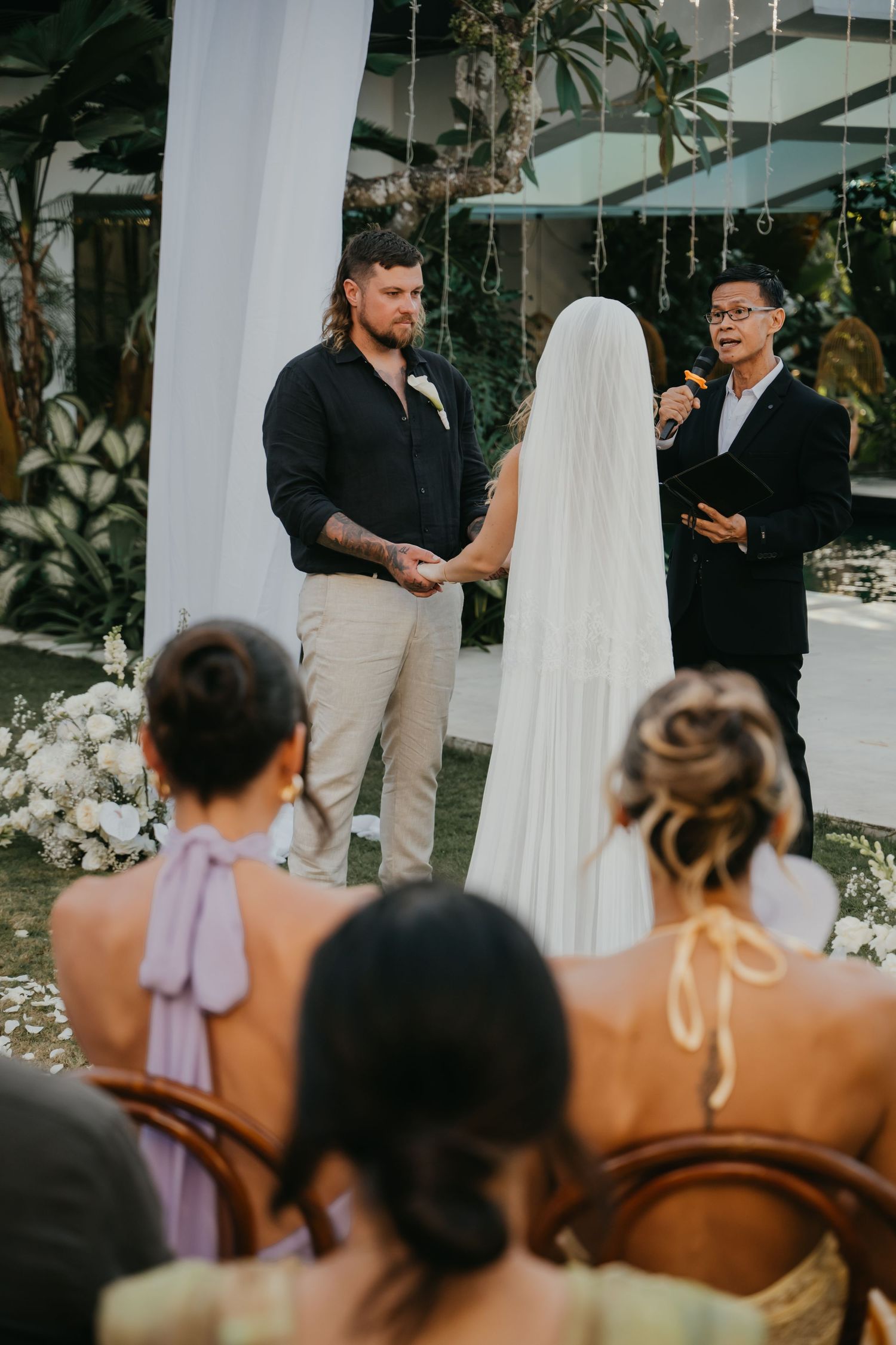 Groom in black shirt holds brides hands as officiant reads vows at outdoor tropical wedding ceremony.
