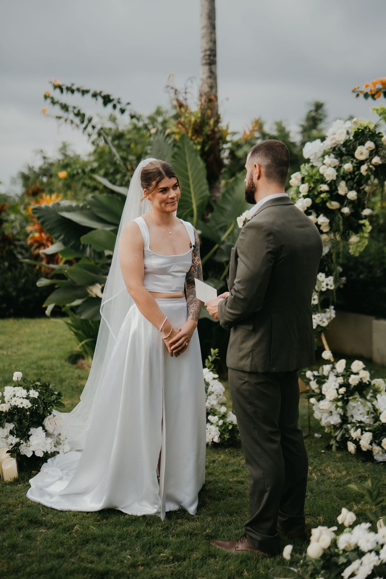 Smiling bride in two-piece white gown holding grooms hands at garden wedding ceremony altar.