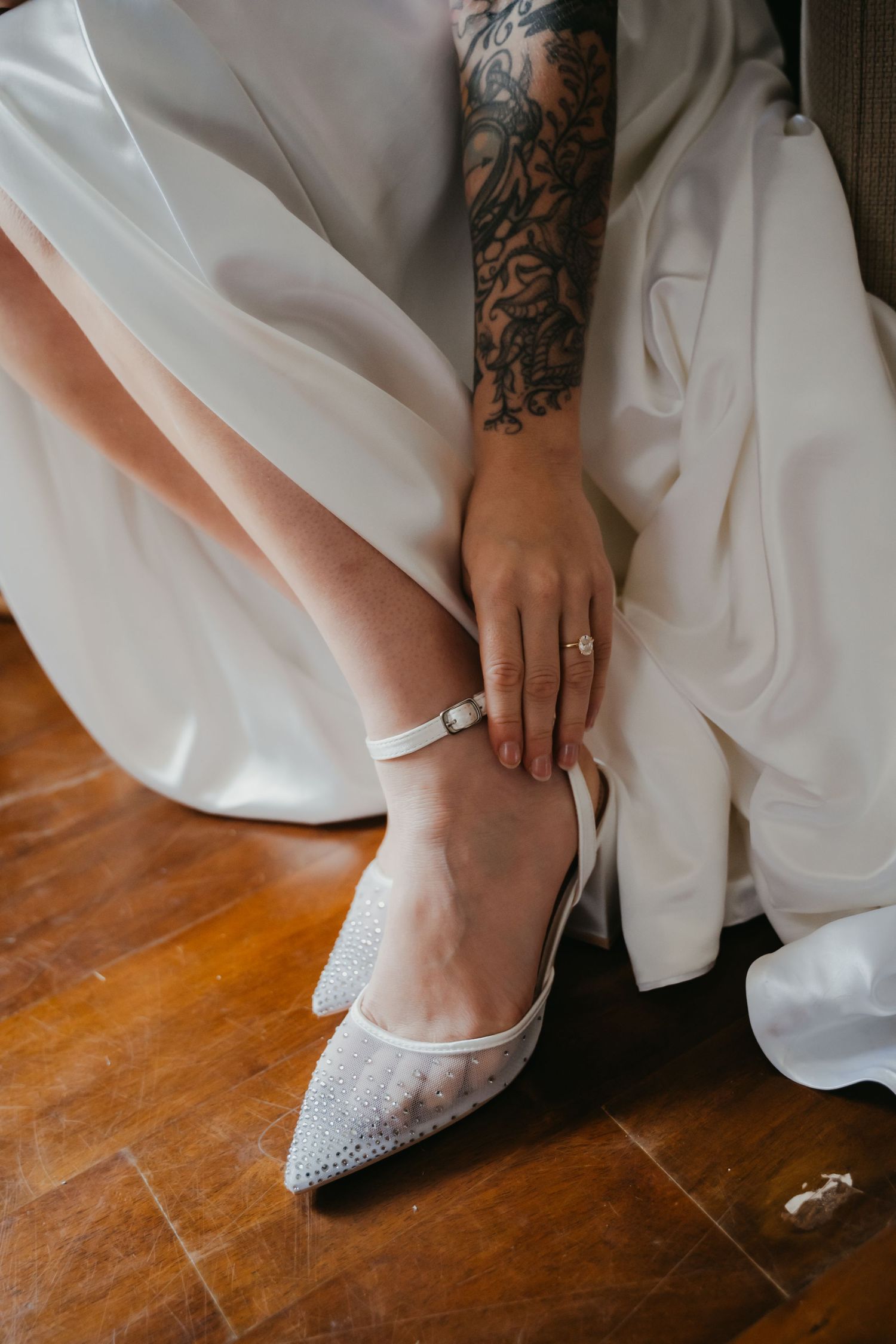 Tattooed bride fastens elegant crystal-embellished white heels with ankle strap on wooden floor in wedding dress.