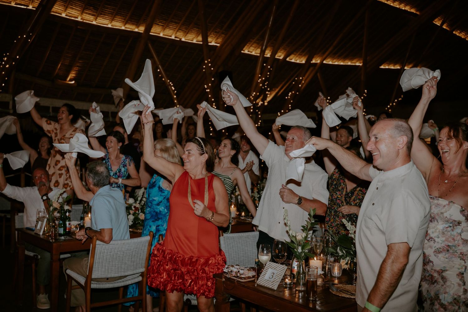 Wedding reception guests dancing and waving napkins in celebration at a rustic venue with warm lighting.