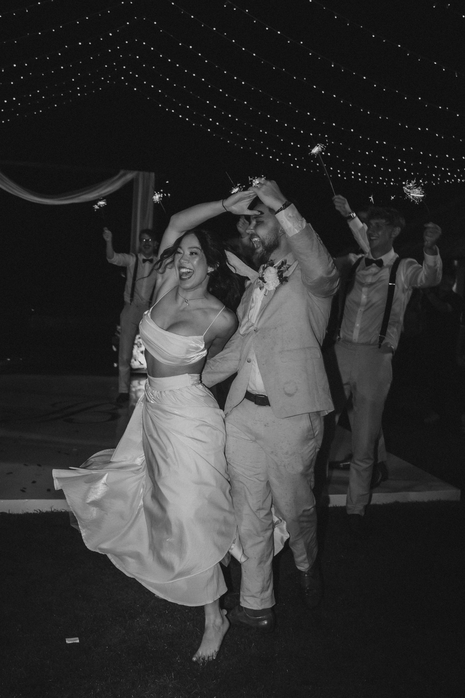 A couple dances joyfully at their wedding reception under twinkling lights, the bride in a flowing white dress and barefoot.