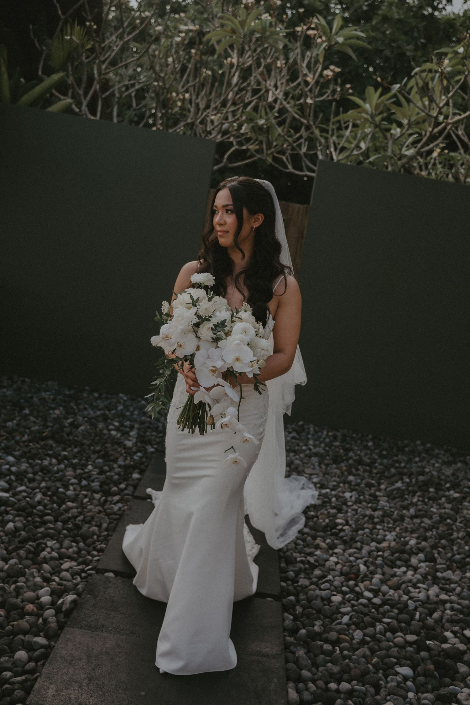 A bride in a sleek white wedding dress holds a bouquet of white orchids while sitting on stone steps in a garden setting.