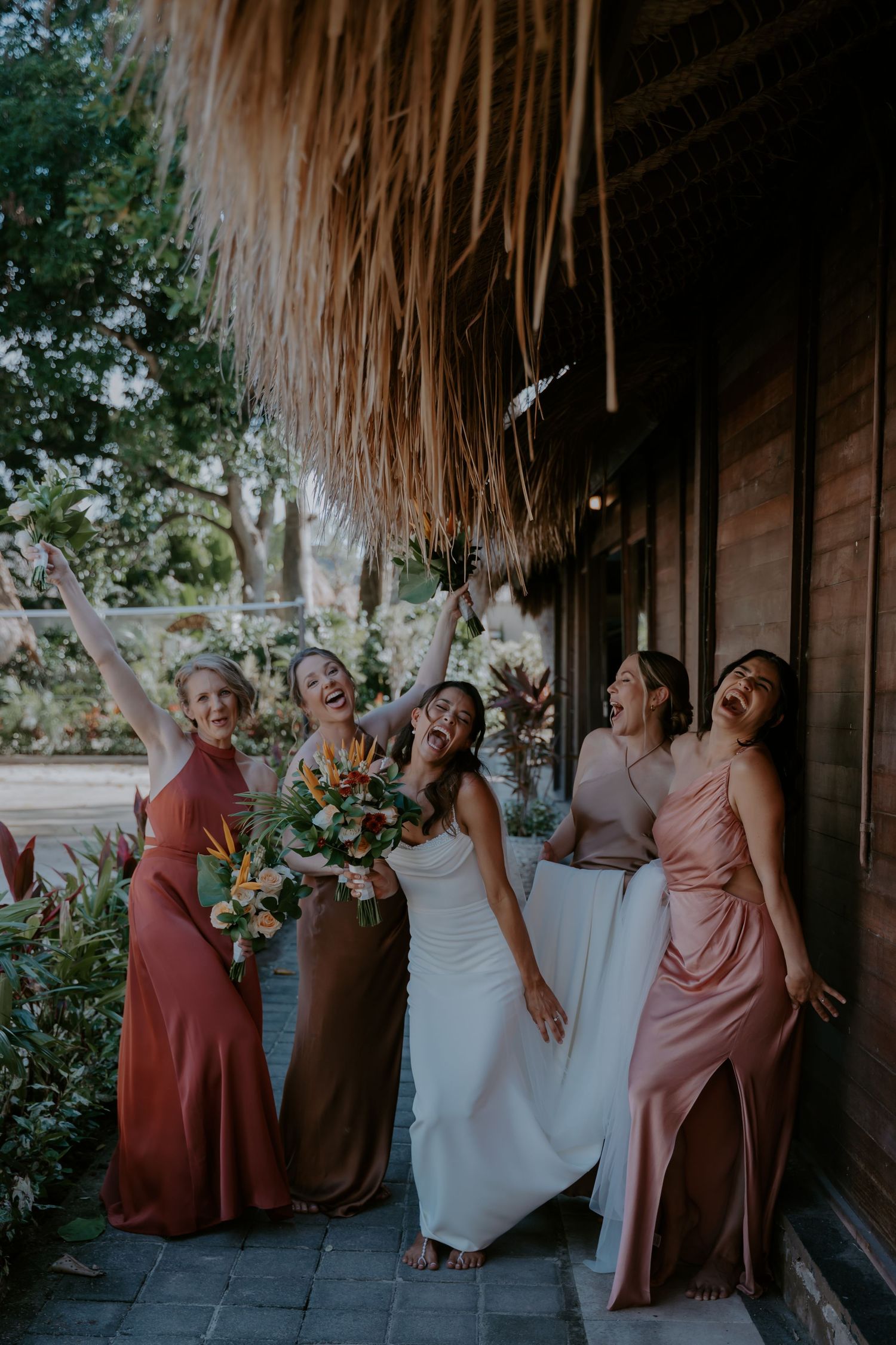 A bridal party in dusty rose and white dresses poses together under a covered walkway with tropical decor.