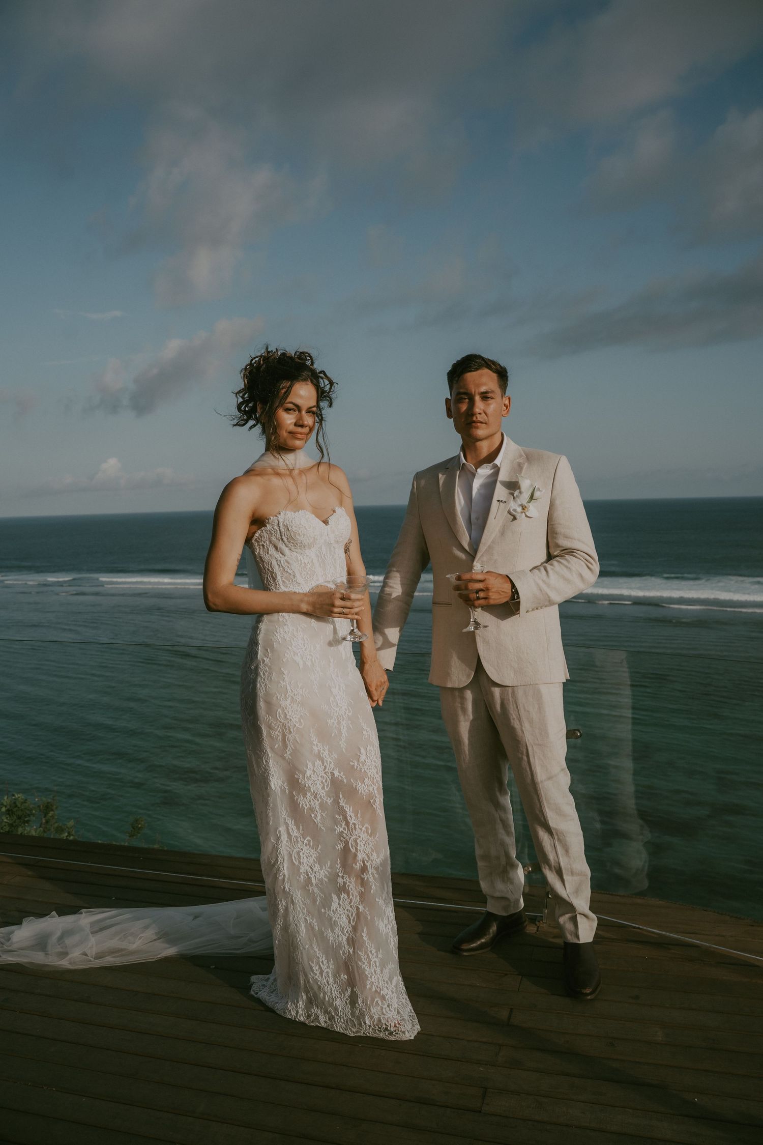 A couple in wedding attire stands on a wooden deck overlooking the ocean during a moody sunset photoshoot.