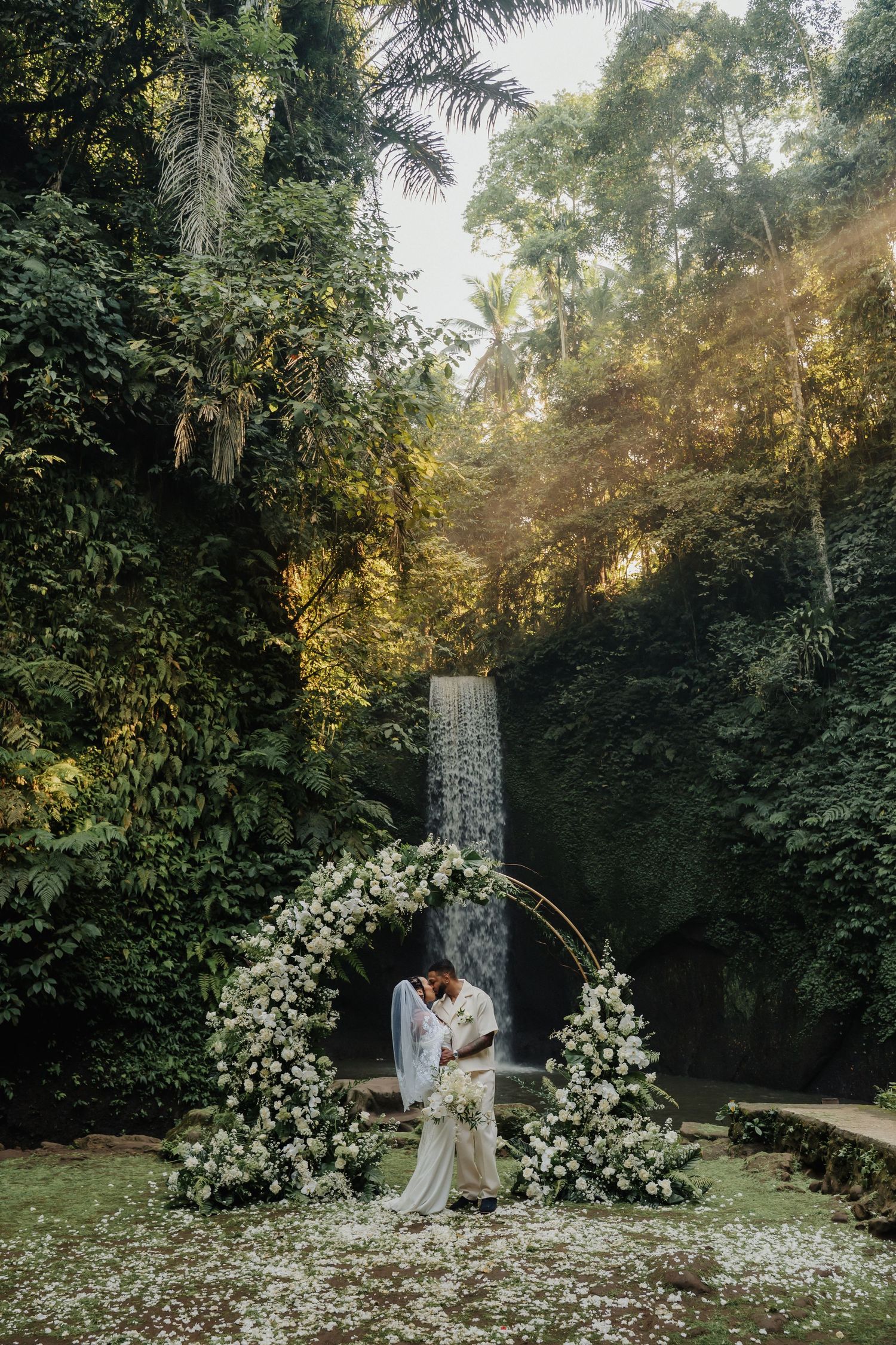 Newlyweds share a romantic kiss beneath a circular white floral arch before a stunning tropical waterfall.