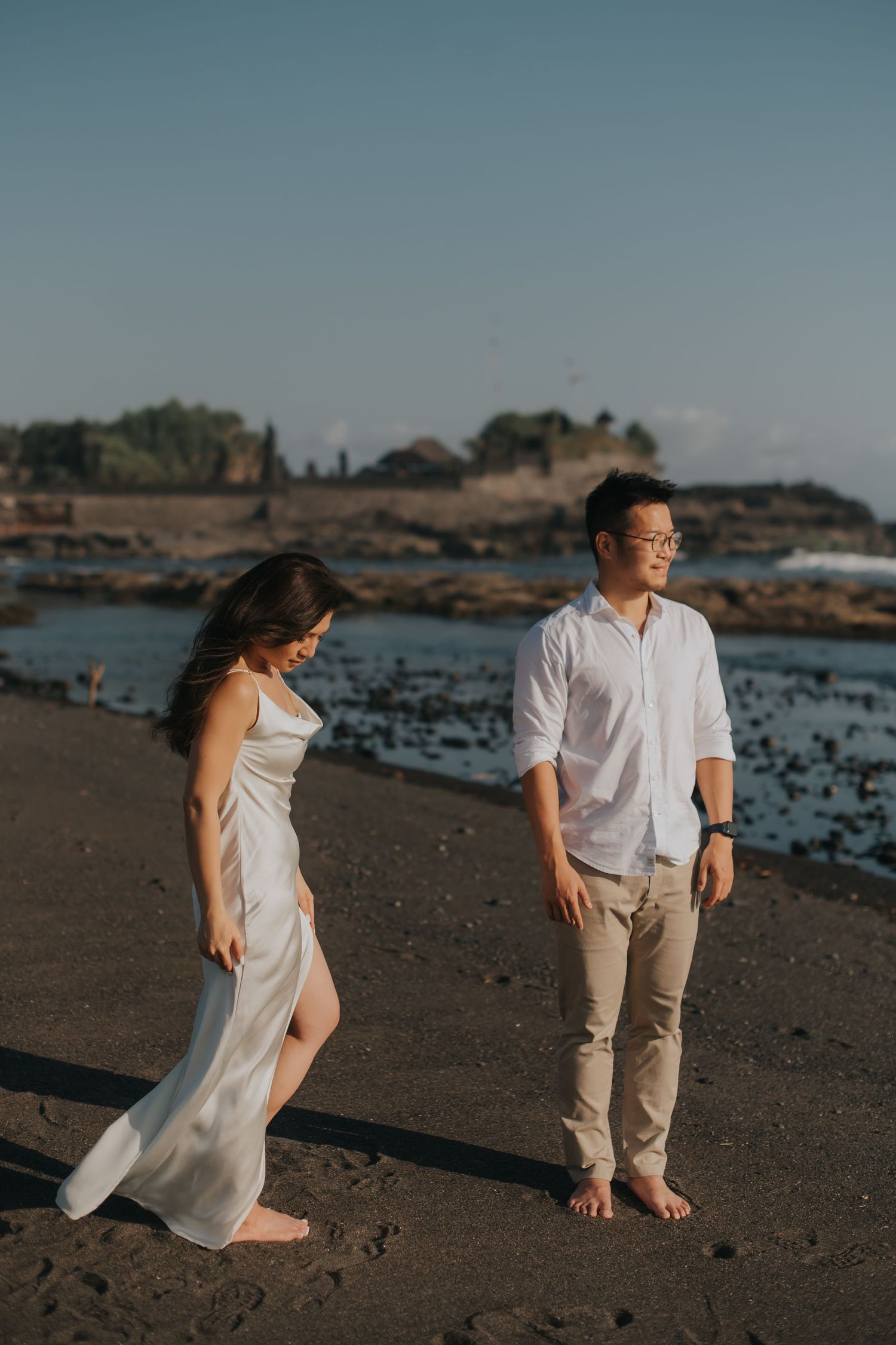Couple in white outfits standing on black sand beach at sunset with rocky coastline and ocean in background.