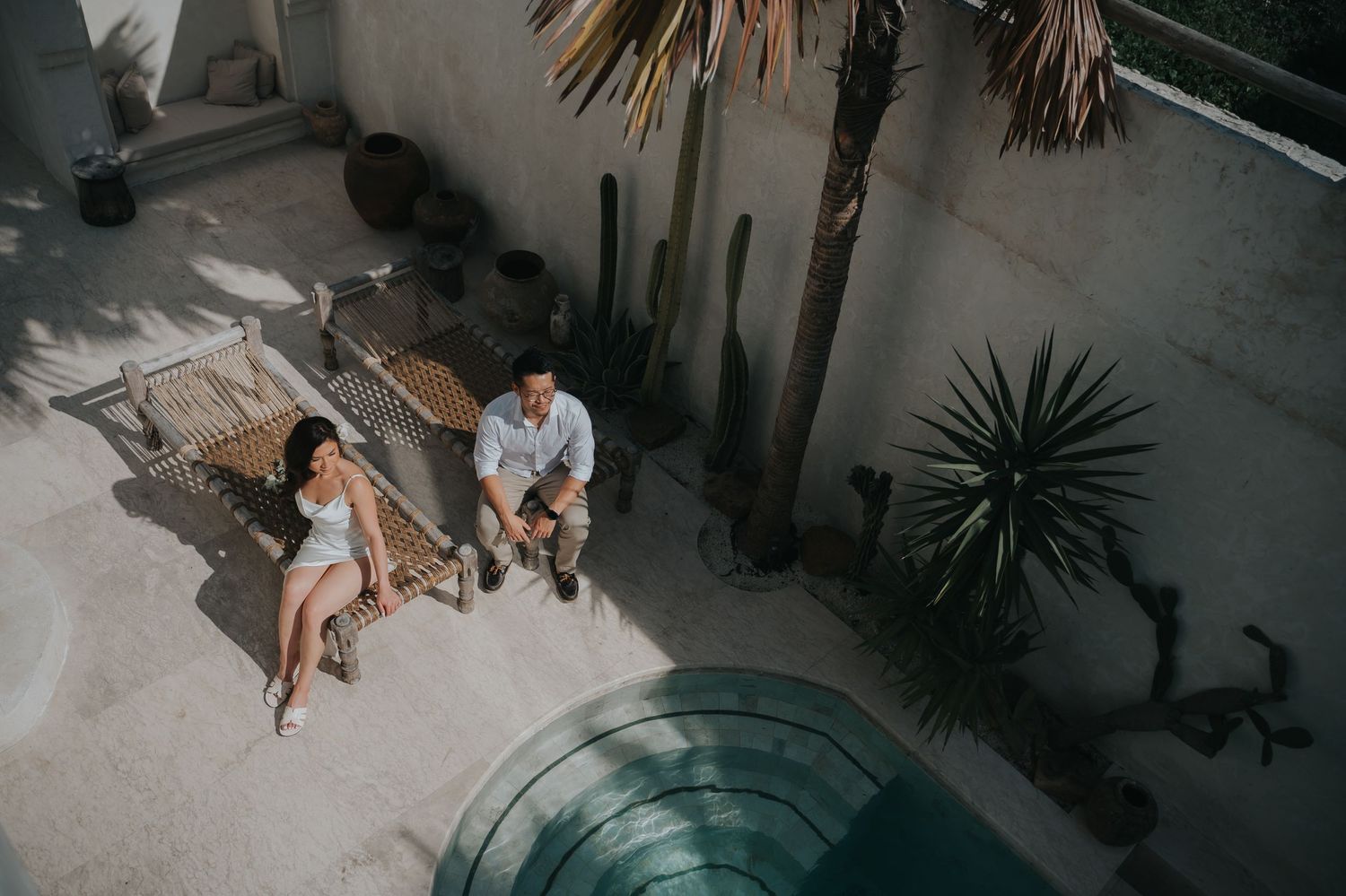 Couple relaxing by a pool patio with cacti and palm trees in a Mediterranean-style courtyard.