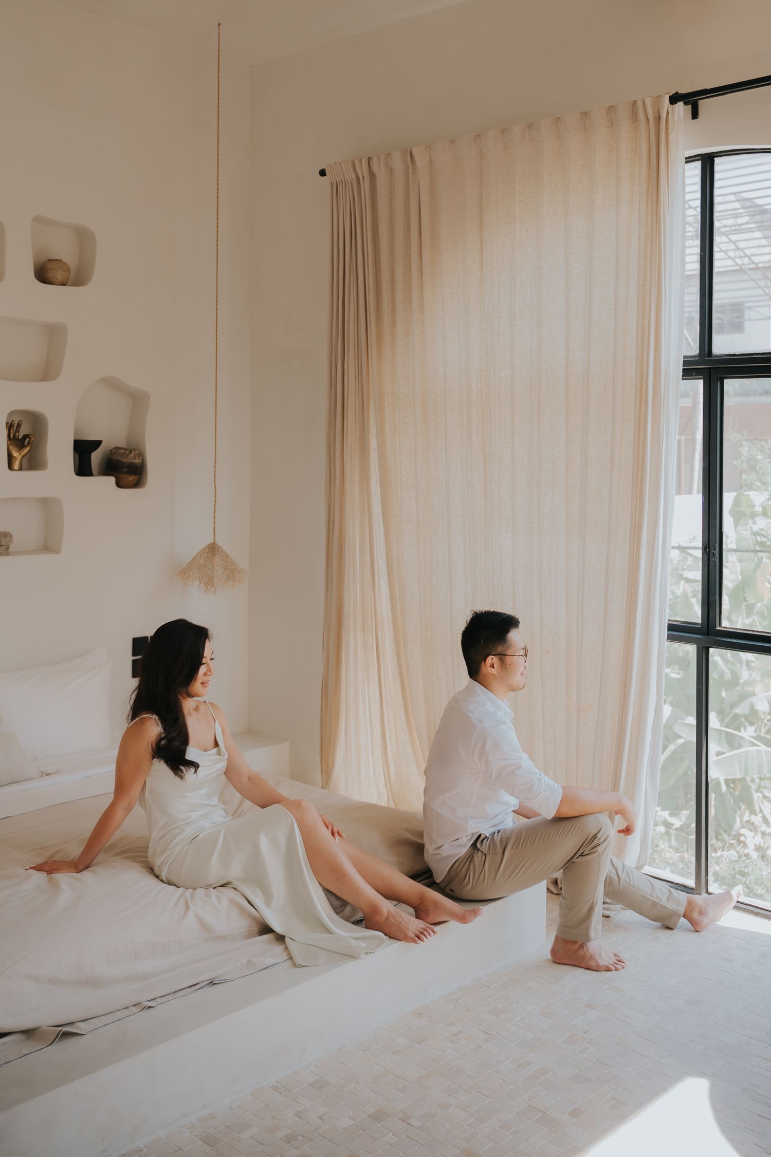 A couple sits back-to-back on a white bed in a minimalist bedroom with neutral tones and sheer curtains.