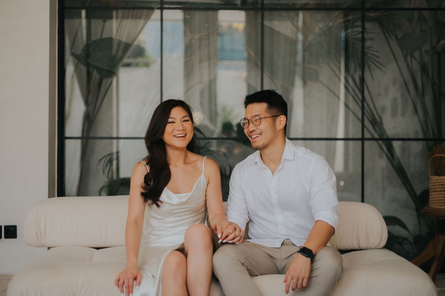 A smiling Asian couple sits together on a white sofa in a modern living room, holding hands in casual elegant attire.