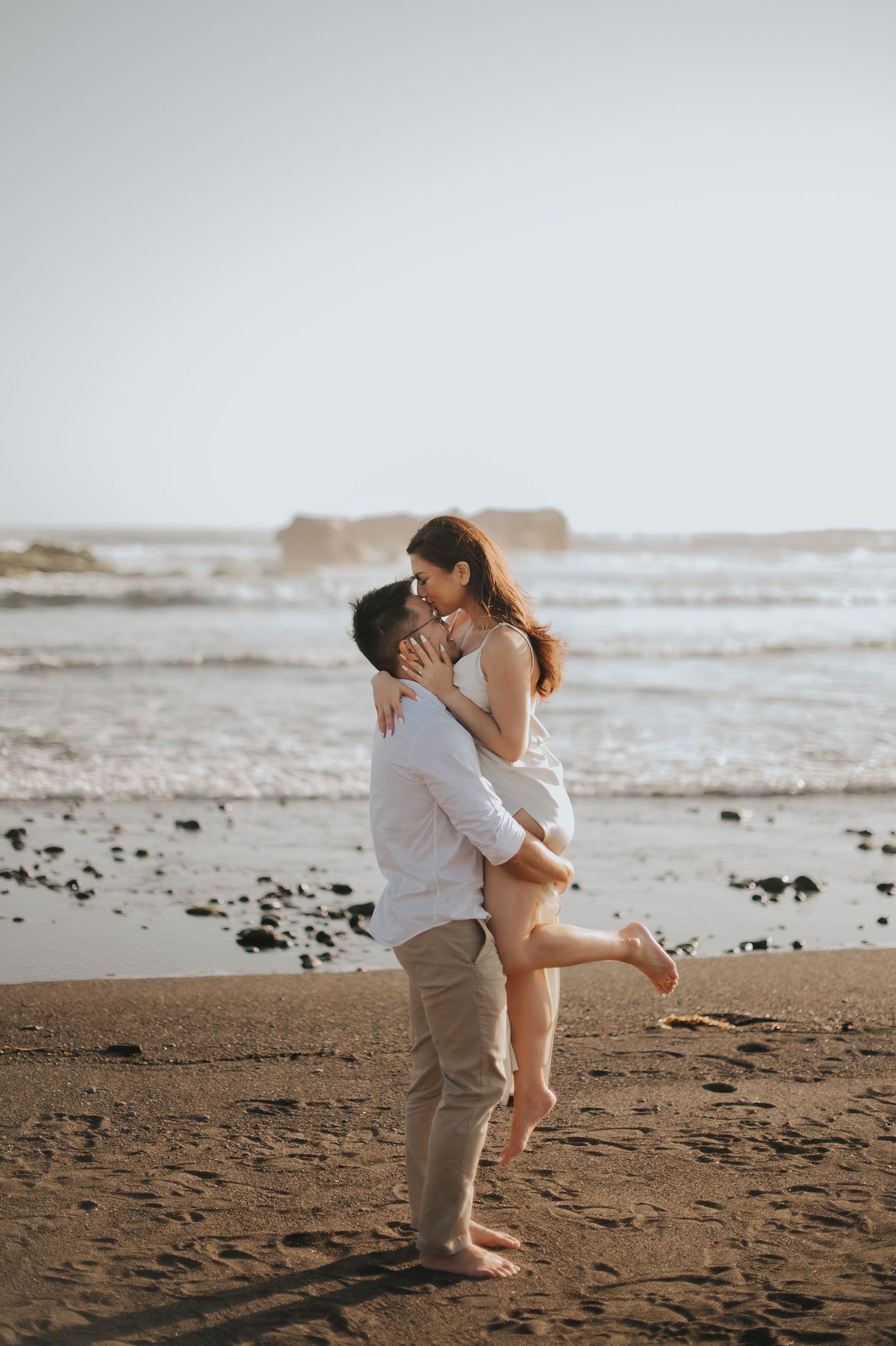 Couple sharing a romantic kiss on a sandy beach at sunset, man lifting woman in white dress near ocean waves.