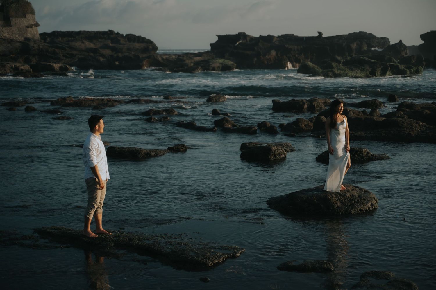 Couple standing on rocky shoreline at dusk with ocean waves, man in white shirt and woman in flowing dress facing each other.