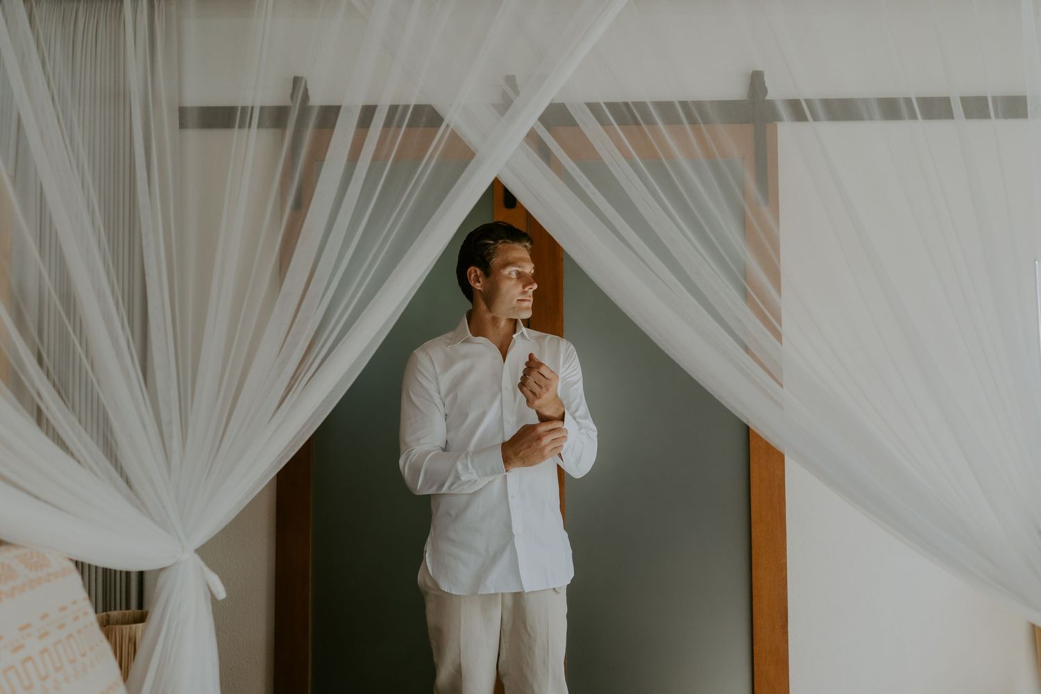 Groom in white shirt adjusting cufflinks standing between sheer white canopy curtains in elegant bedroom.