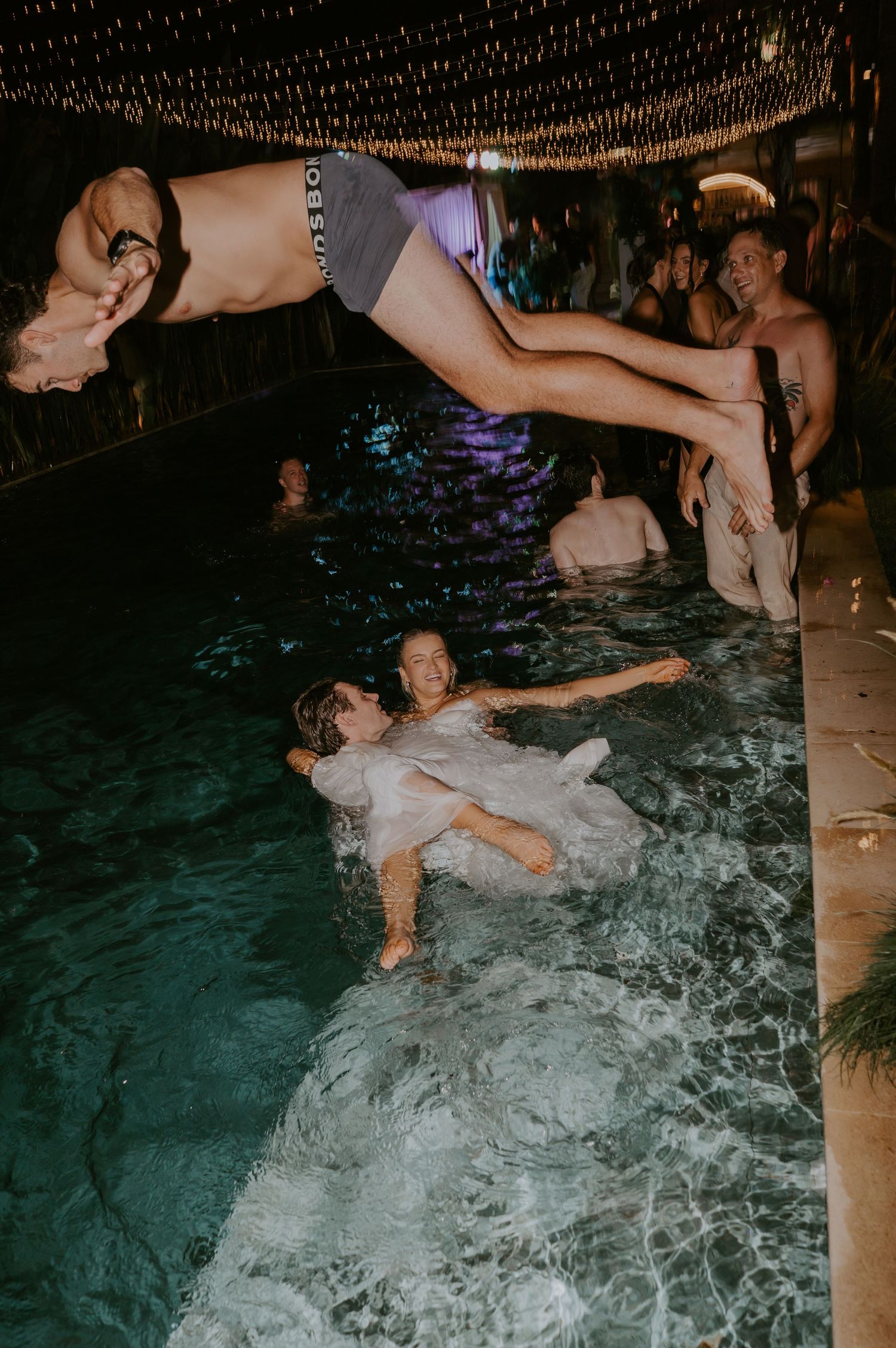 Wedding guest dives into pool as bride floats in water during lively late-night celebration.