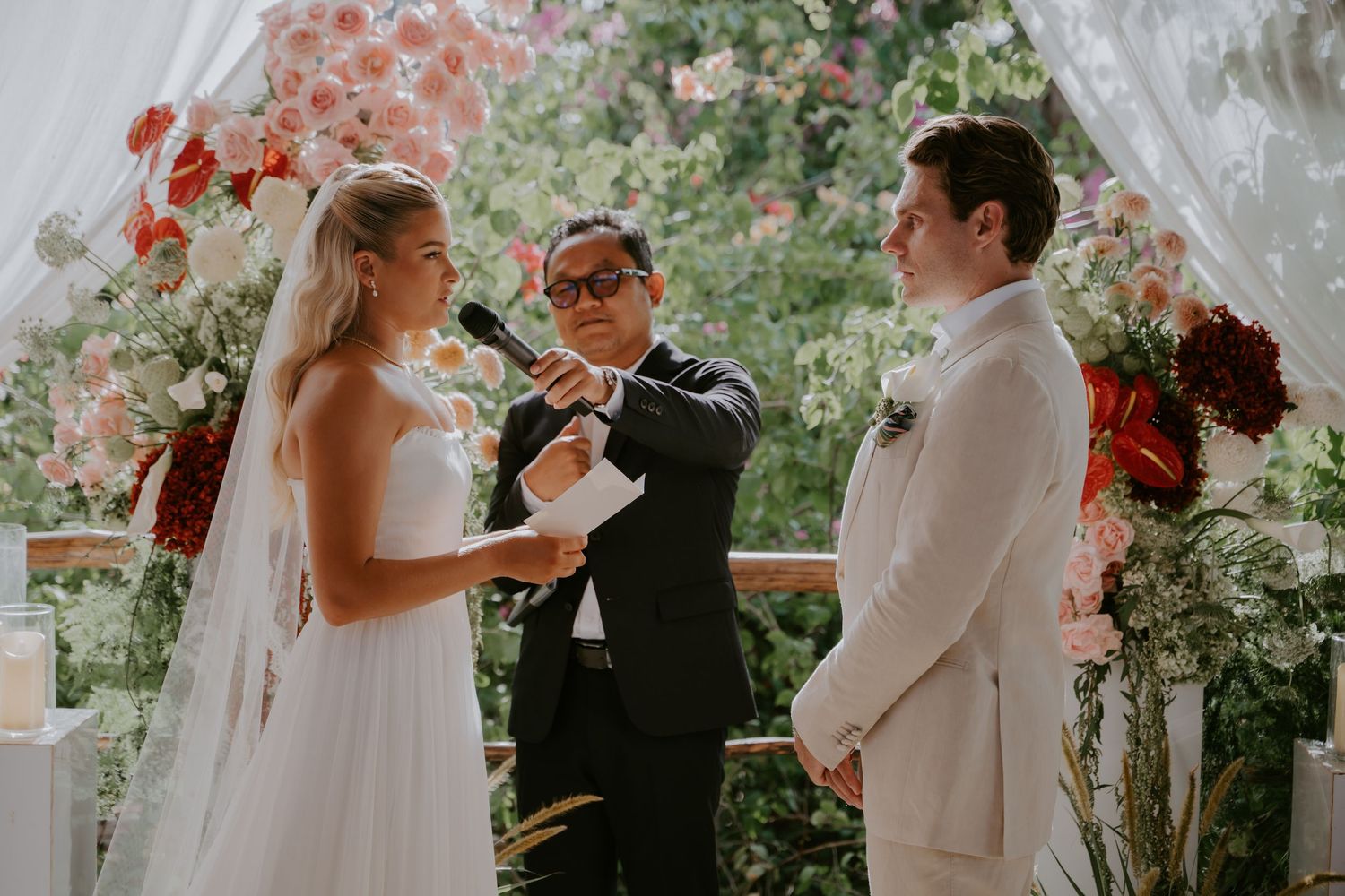 Bride reading vows to groom at floral altar with officiant during outdoor wedding ceremony.