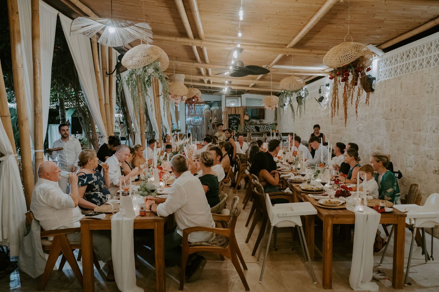 Wedding guests enjoy dinner at long tables in a beautifully decorated tropical venue.