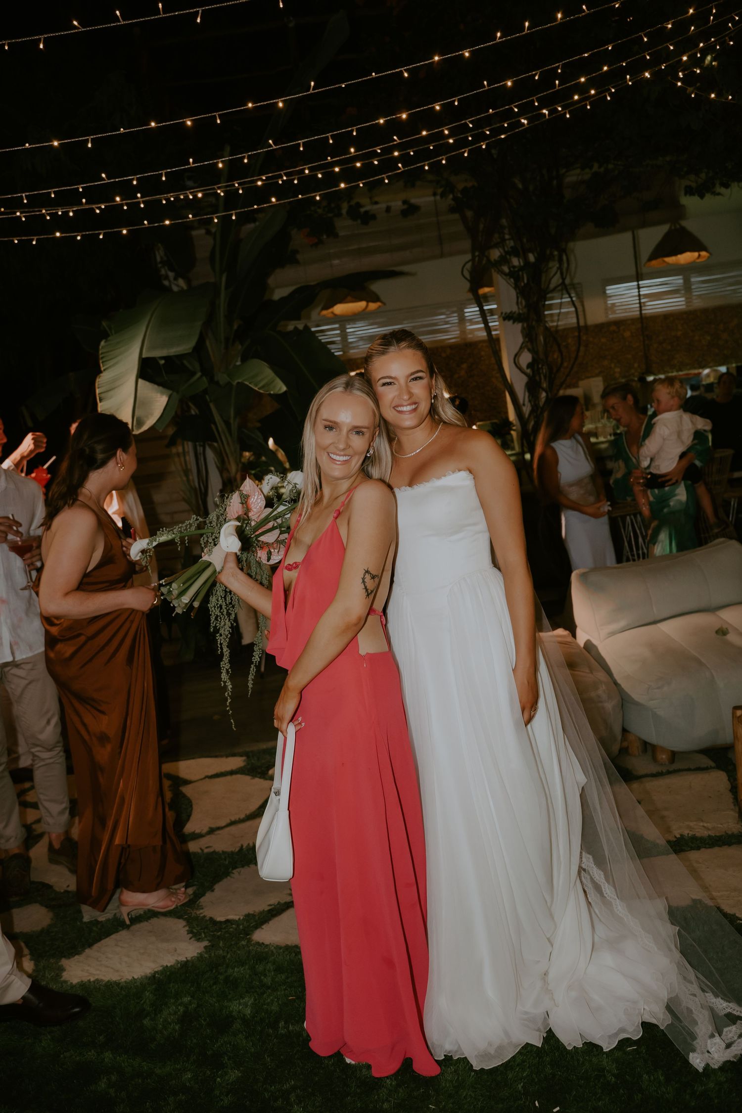 Bride in white strapless gown poses with guest in coral dress at evening reception.