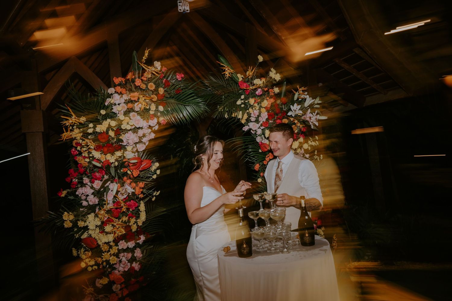 Newlyweds cutting wedding cake beside colorful tropical floral arch during evening Bali reception celebration.