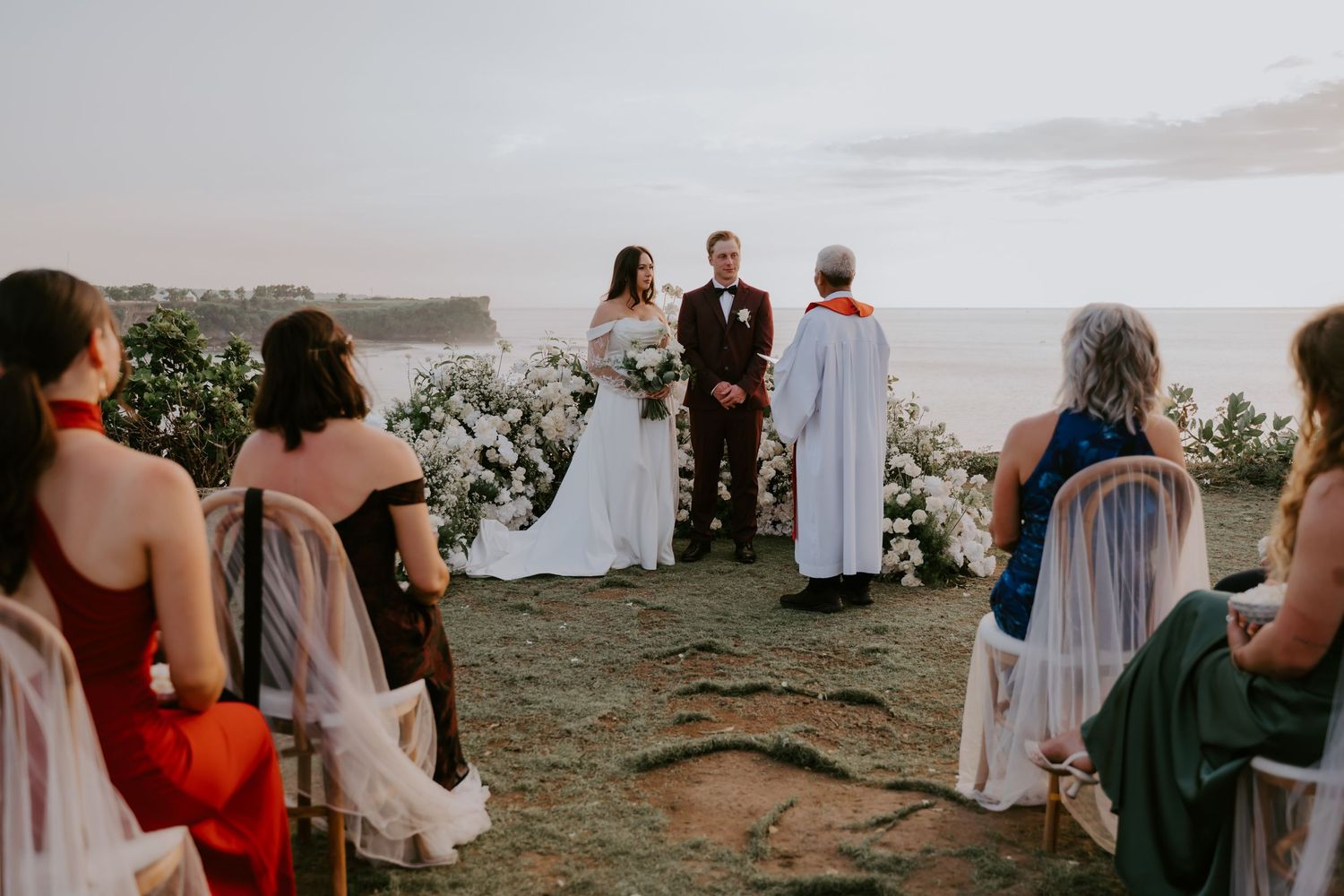 Outdoor cliff-top wedding ceremony with couple exchanging vows before an ocean backdrop at sunset.