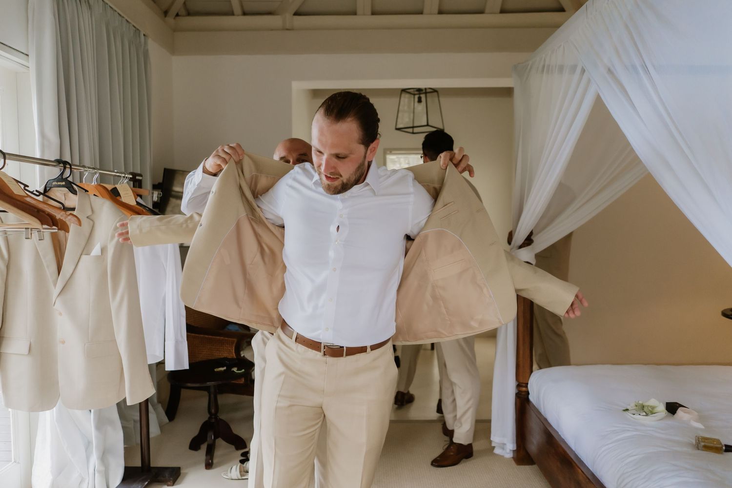 Groom trying on a beige wedding suit jacket in a bright room with suits hanging nearby.