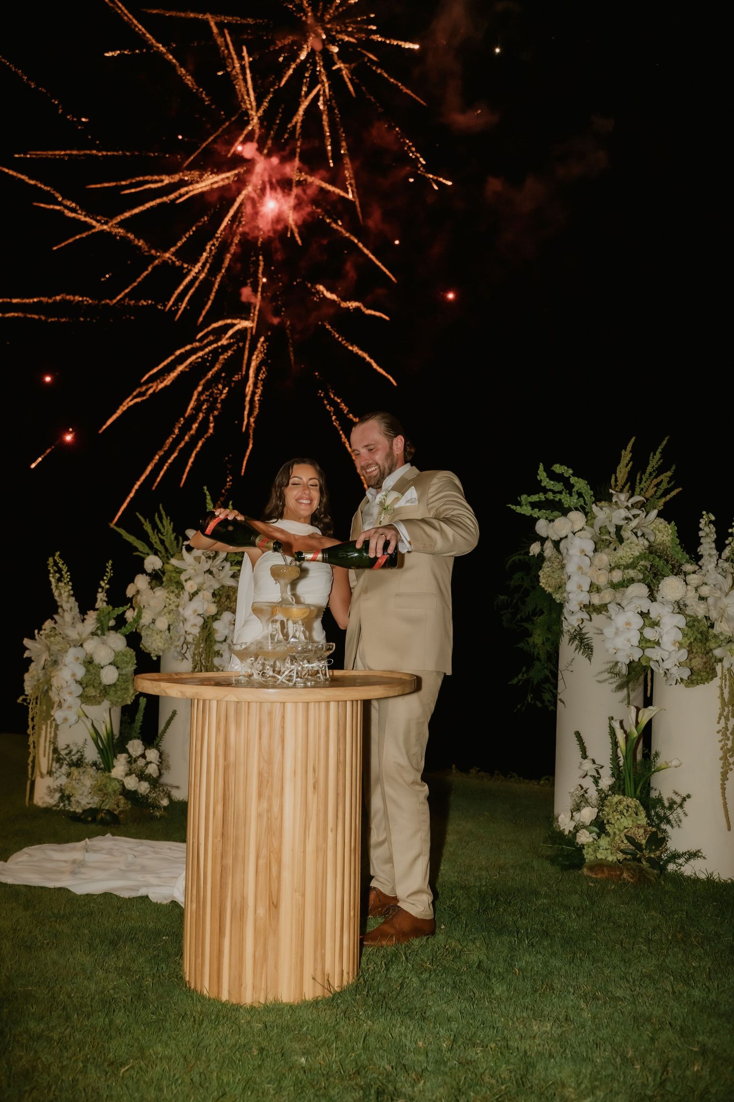 Newlyweds pour champagne at wooden table with stunning fireworks display and white floral arrangements.