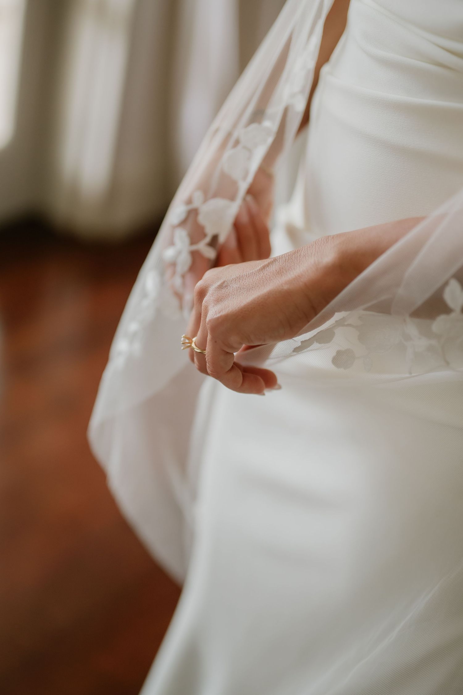 Close-up of bride in white lace wedding dress holding floral veil with gold ring on finger.