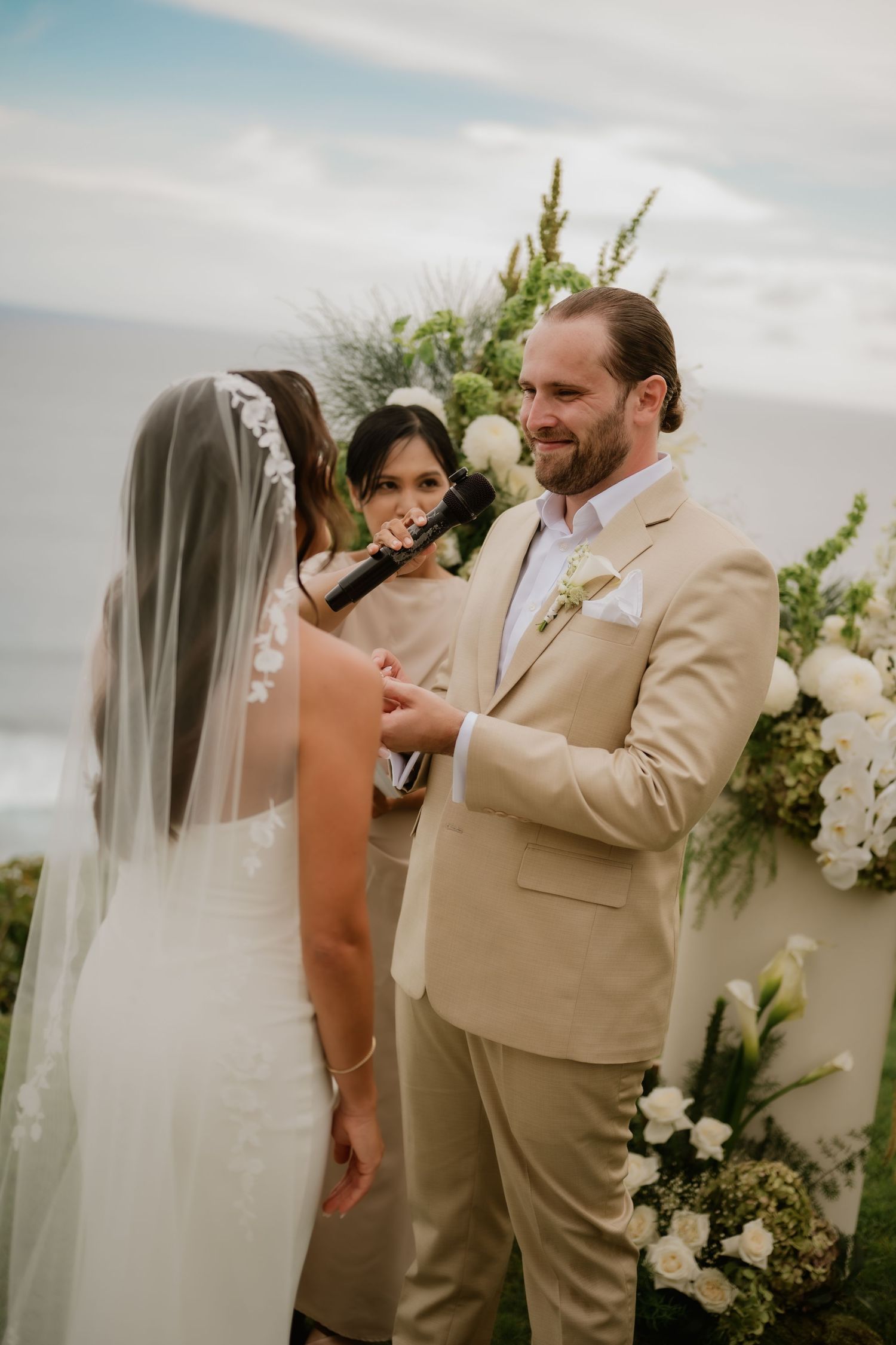 Groom in tan suit smiles at bride during outdoor oceanfront wedding ceremony with white floral arch.