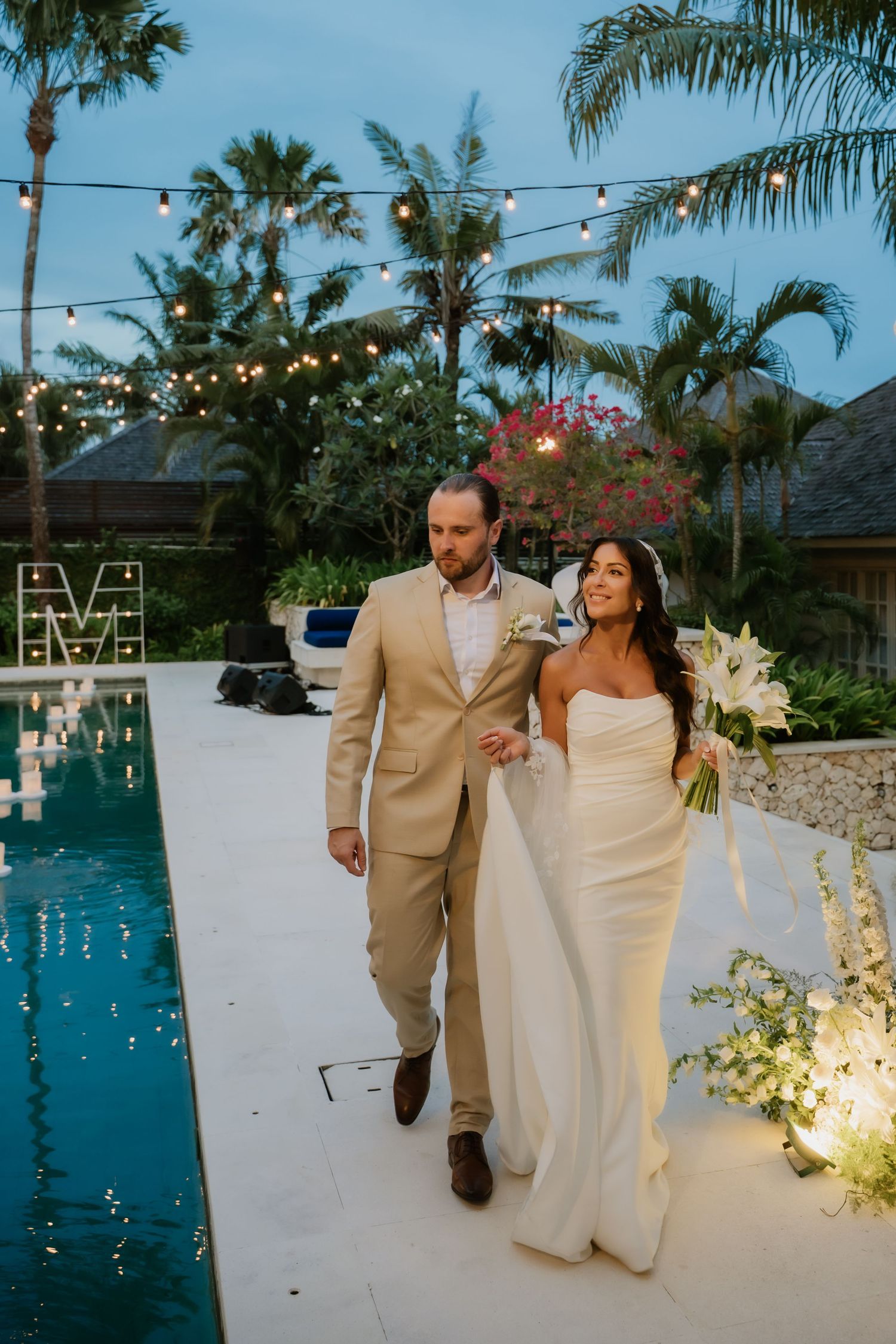 Elegant couple walks poolside at dusk during tropical wedding reception with string lights and palm trees.