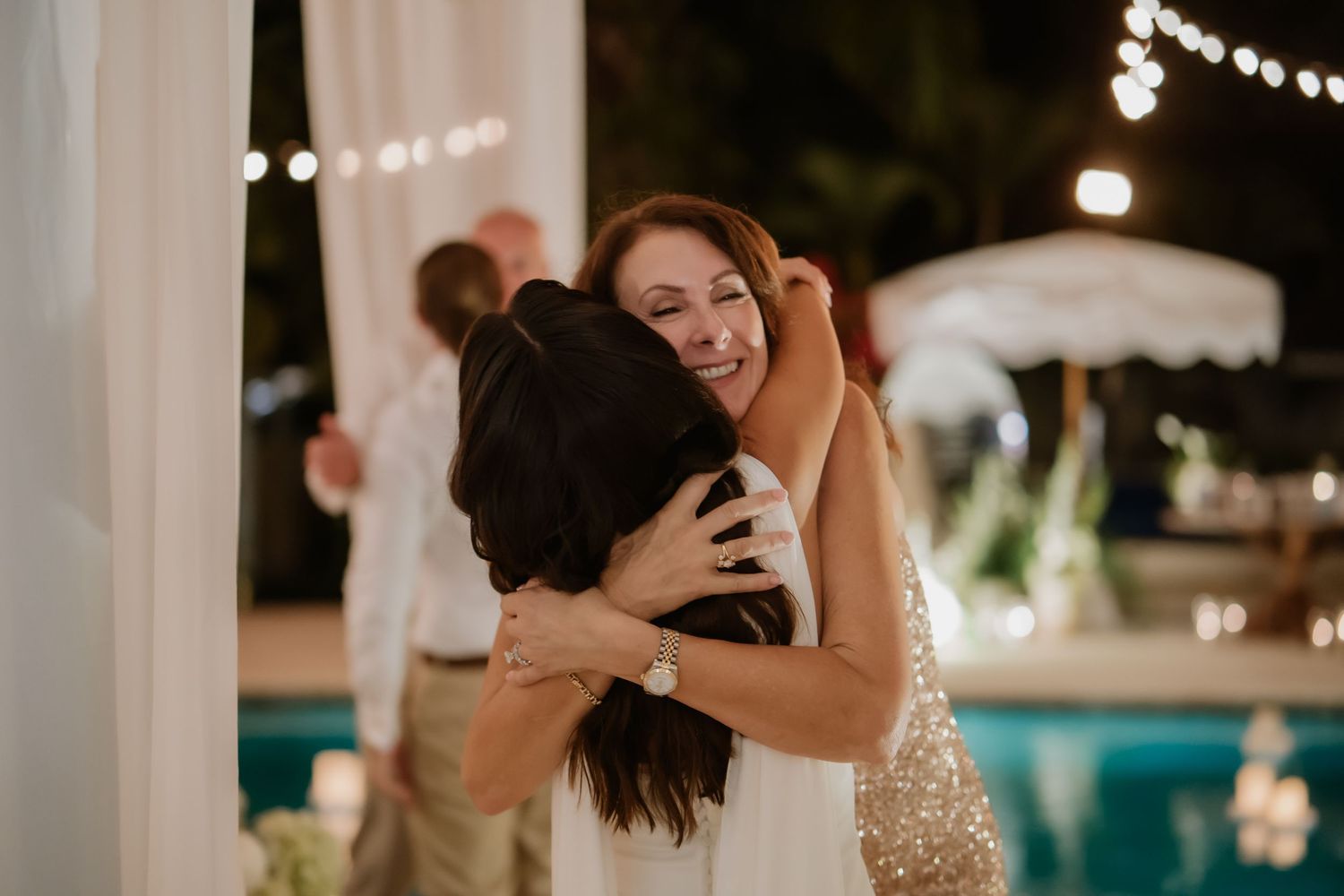 Bride embraces a smiling guest in a gold sequin dress at an outdoor evening wedding reception.