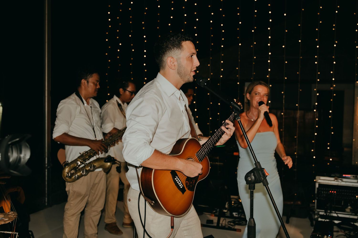 Live wedding band performs with guitarist and female vocalist under warm fairy lights at an outdoor reception.