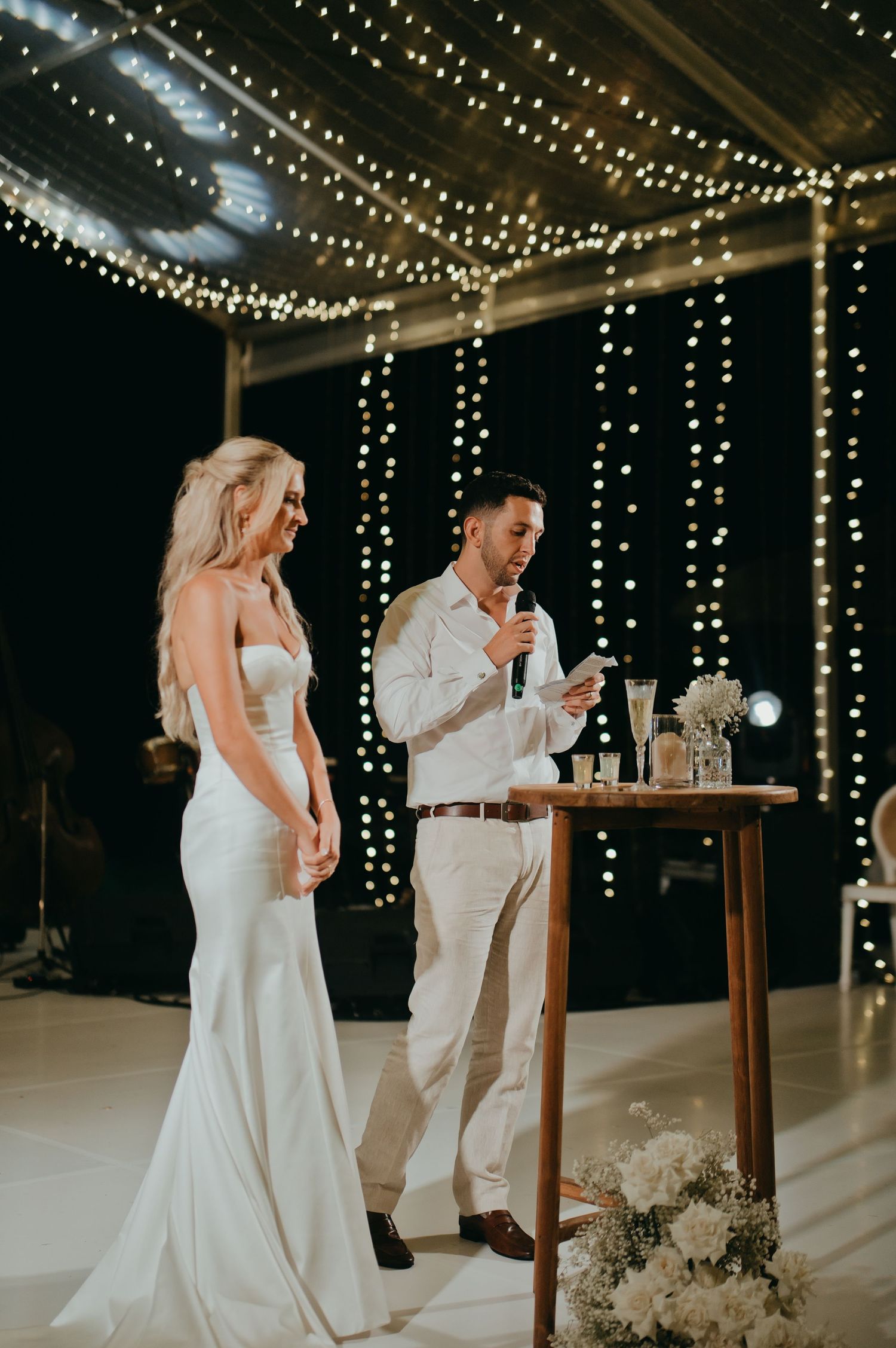 Bride and groom giving a speech under fairy lights at their romantic evening wedding reception.