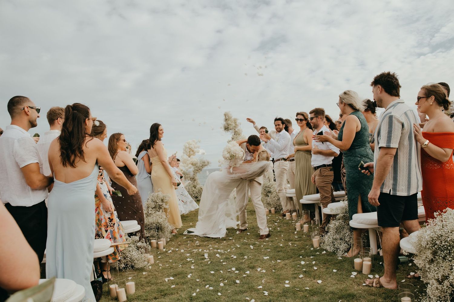 Wedding guests toss flower petals as the bride and groom walk back down the aisle at an outdoor ceremony.