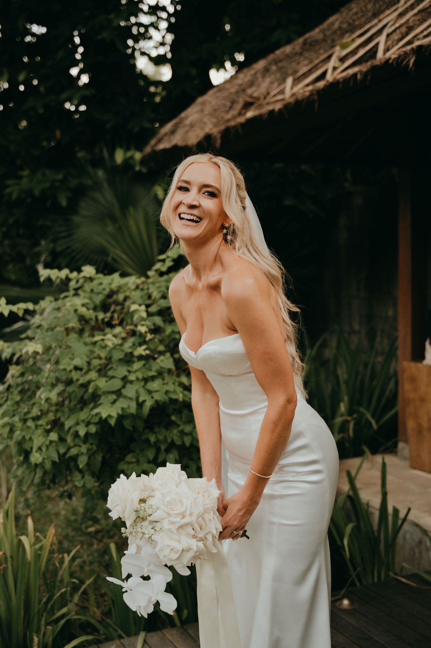 Radiant blonde bride in strapless white satin gown laughing while holding white floral bouquet in tropical setting.