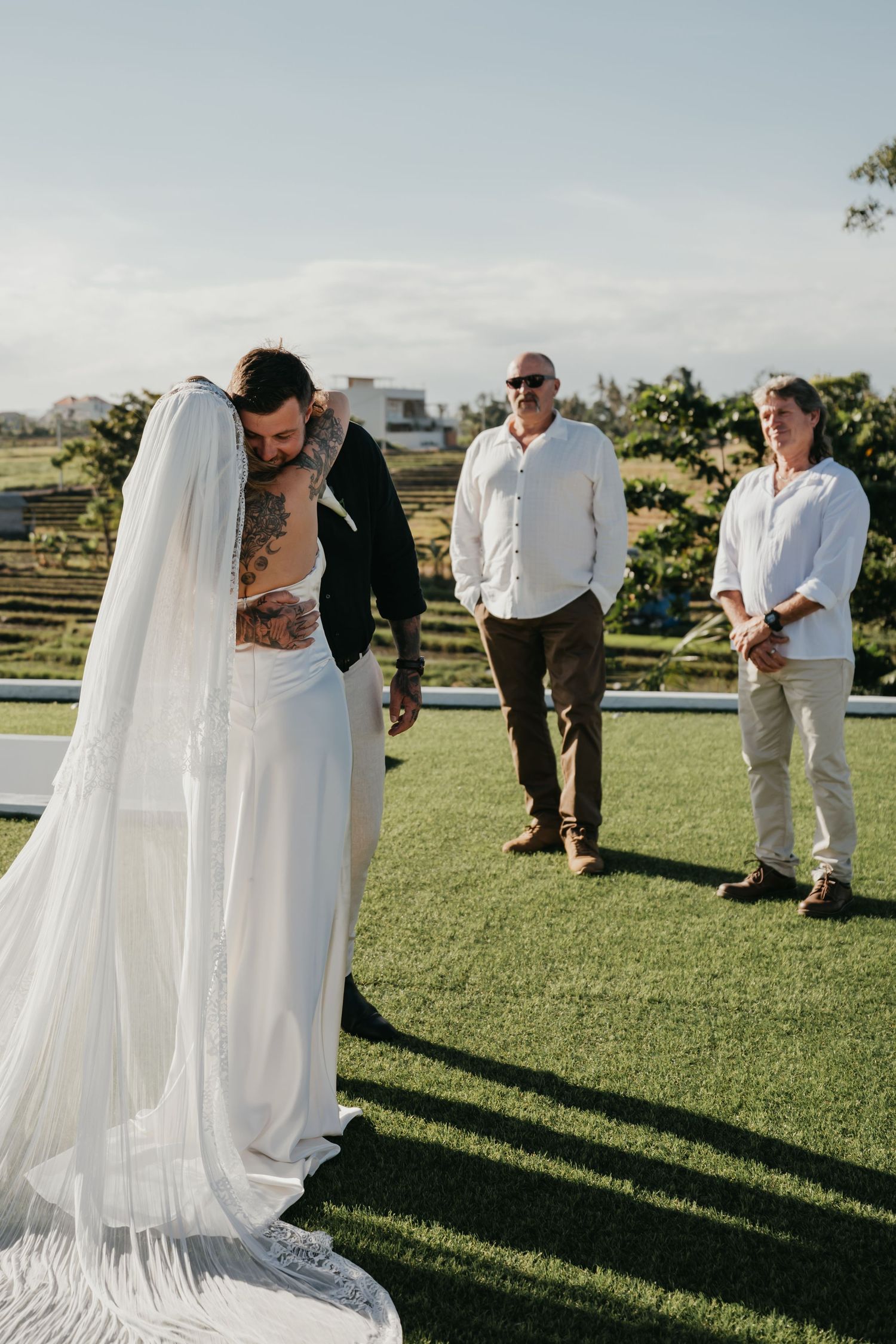 Groom with tattoos embraces bride in white wedding dress and veil during outdoor ceremony.