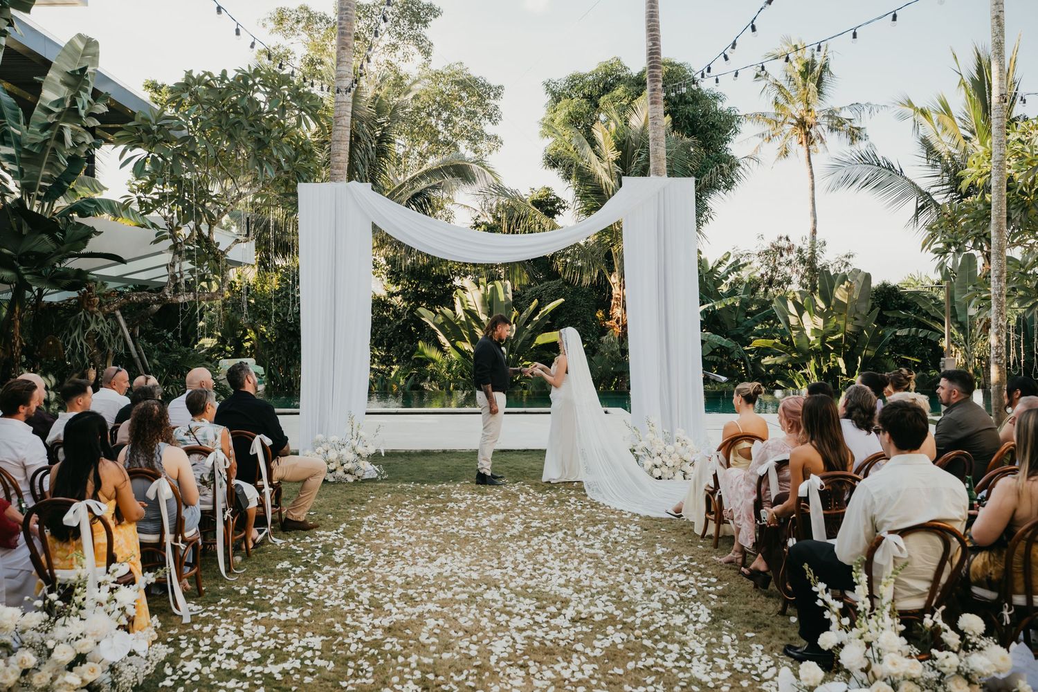 Elegant outdoor tropical wedding ceremony with white petal aisle and draped arch at sunset.