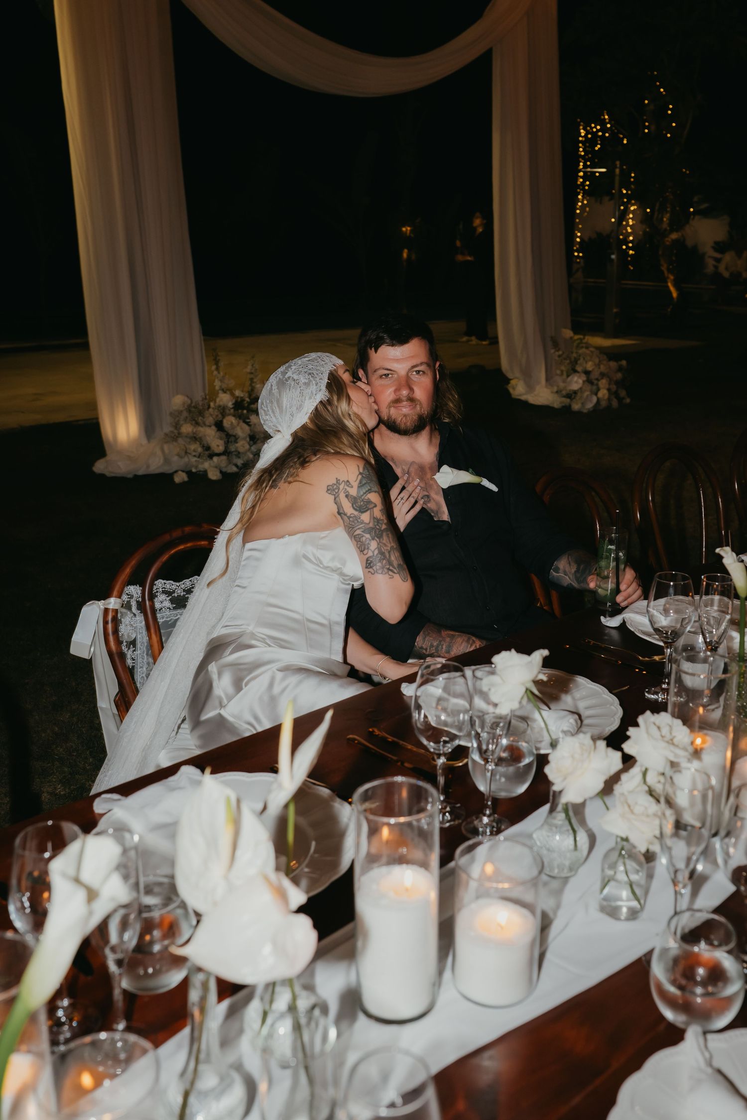 Bride and groom sitting at wedding reception table at night with floral centerpieces.