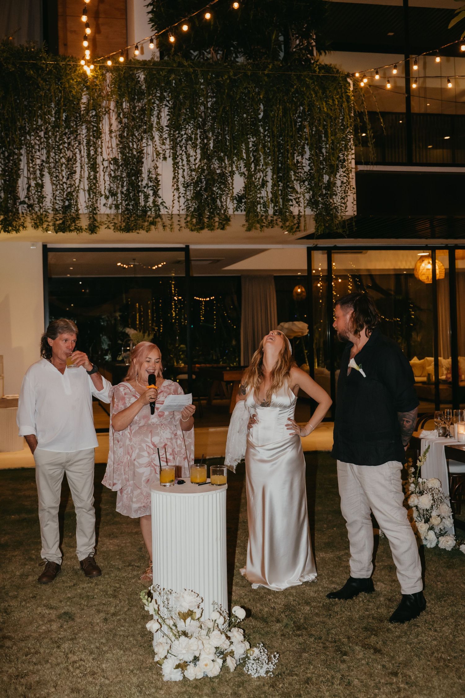Wedding party laughs during outdoor evening reception speeches near a white pedestal with floral arrangements.