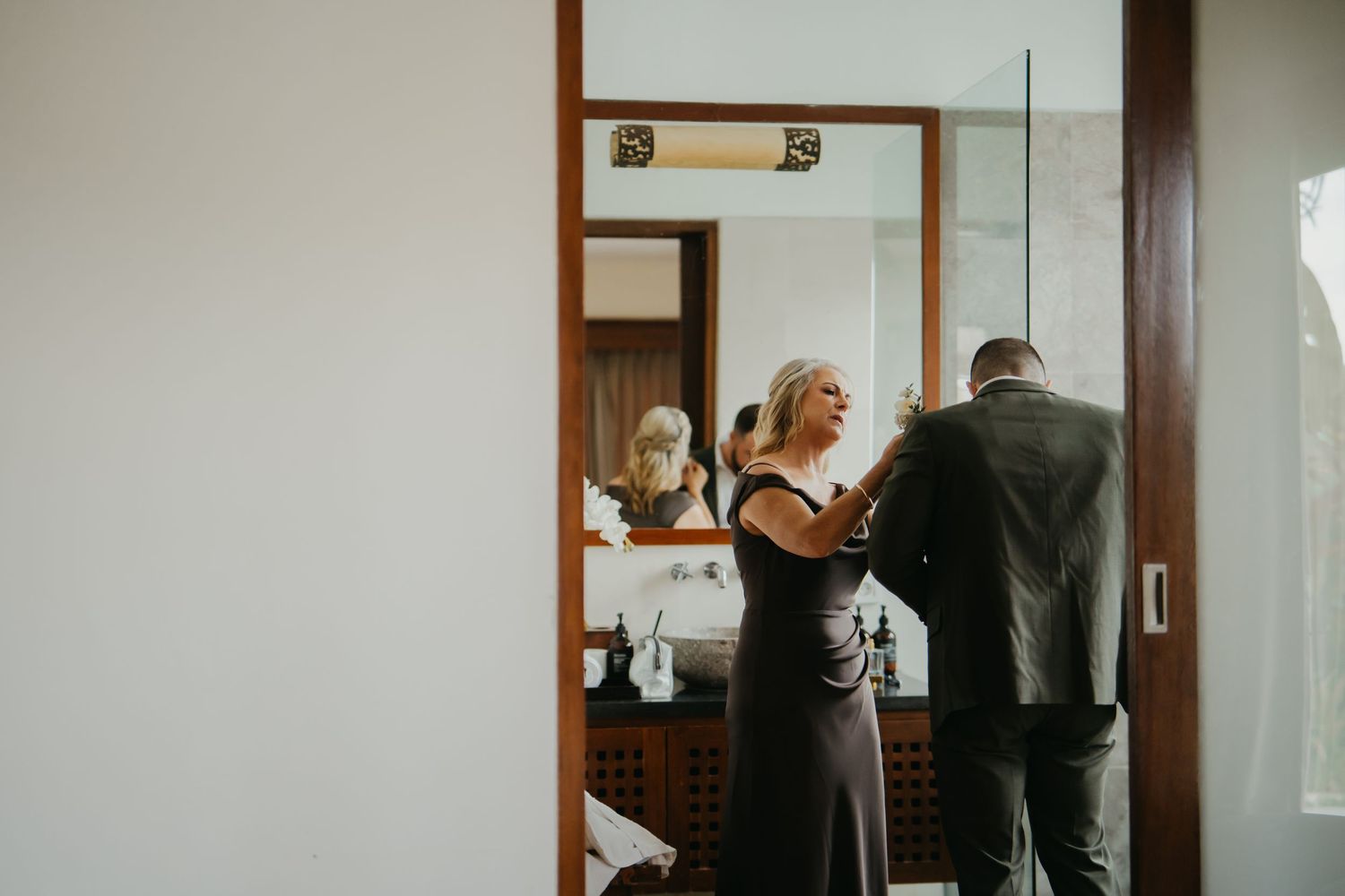 A woman in a dark gown helps a groom with his suit jacket in an elegant bathroom mirror reflection.