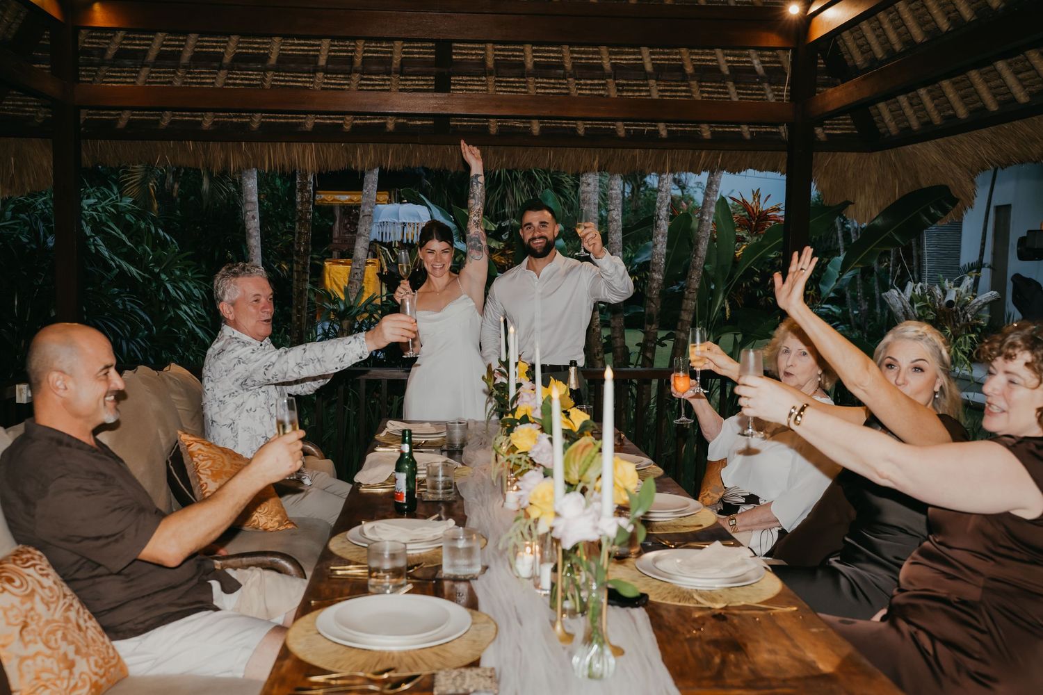 Wedding party raises glasses in a toast at candlelit dinner table in tropical pavilion.