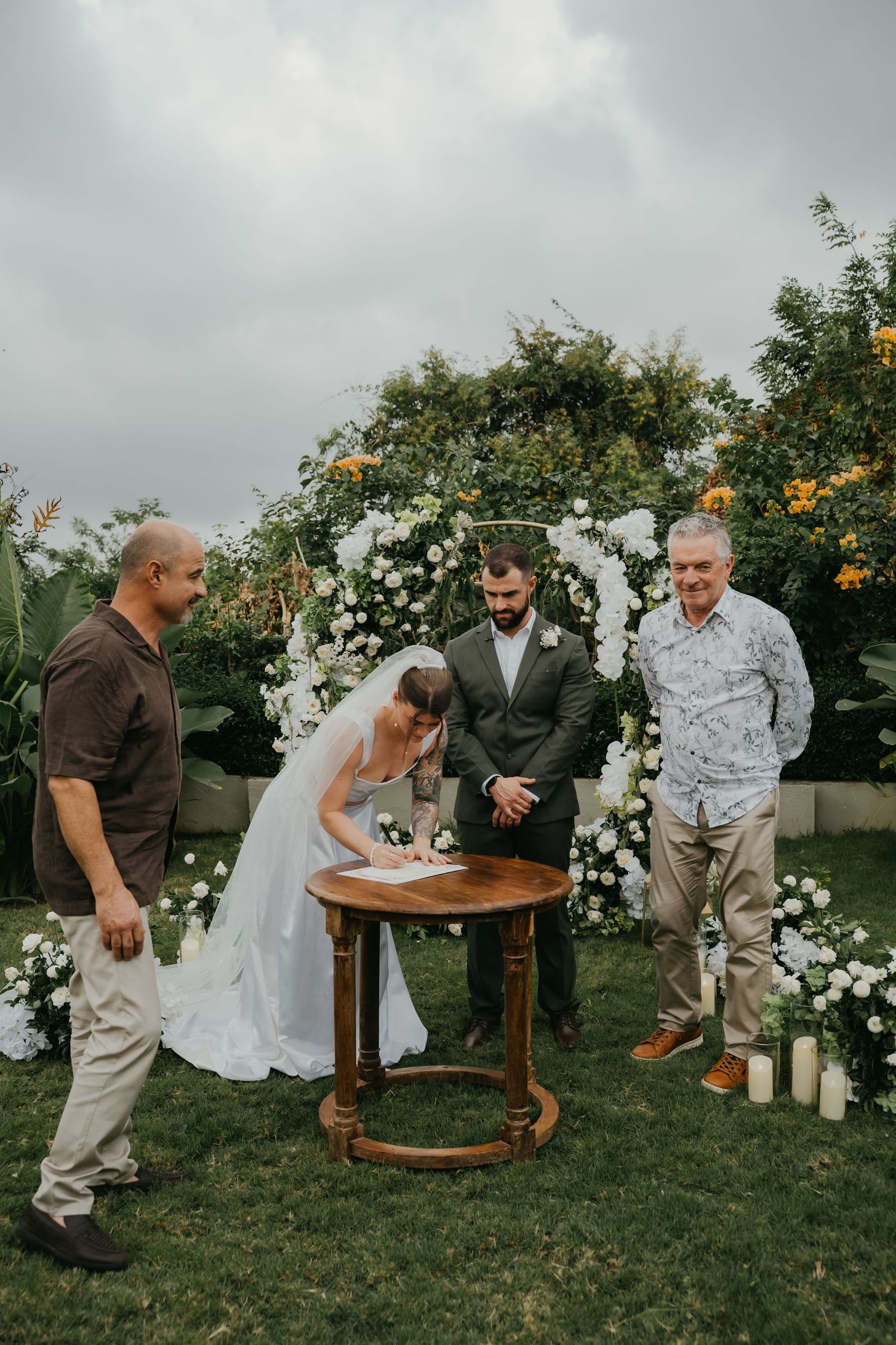 Bride signs wedding register at wooden table with groom and witnesses under white floral arch outdoors.